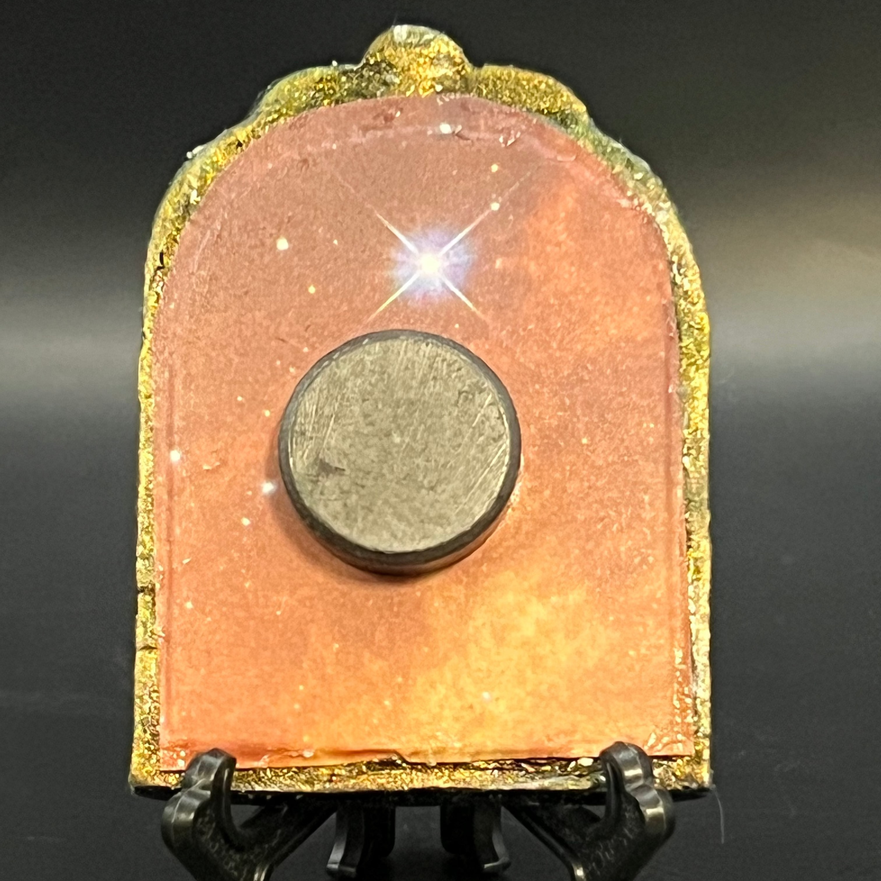 This is the handmade Alignment Miniature Painting by Chris Foster Design. This is showcasing the back. Decorative piece with a colorful, orange/yellow design with glistening stars on a stand against a dark background. There is a magnet and against a miniature picture frame.