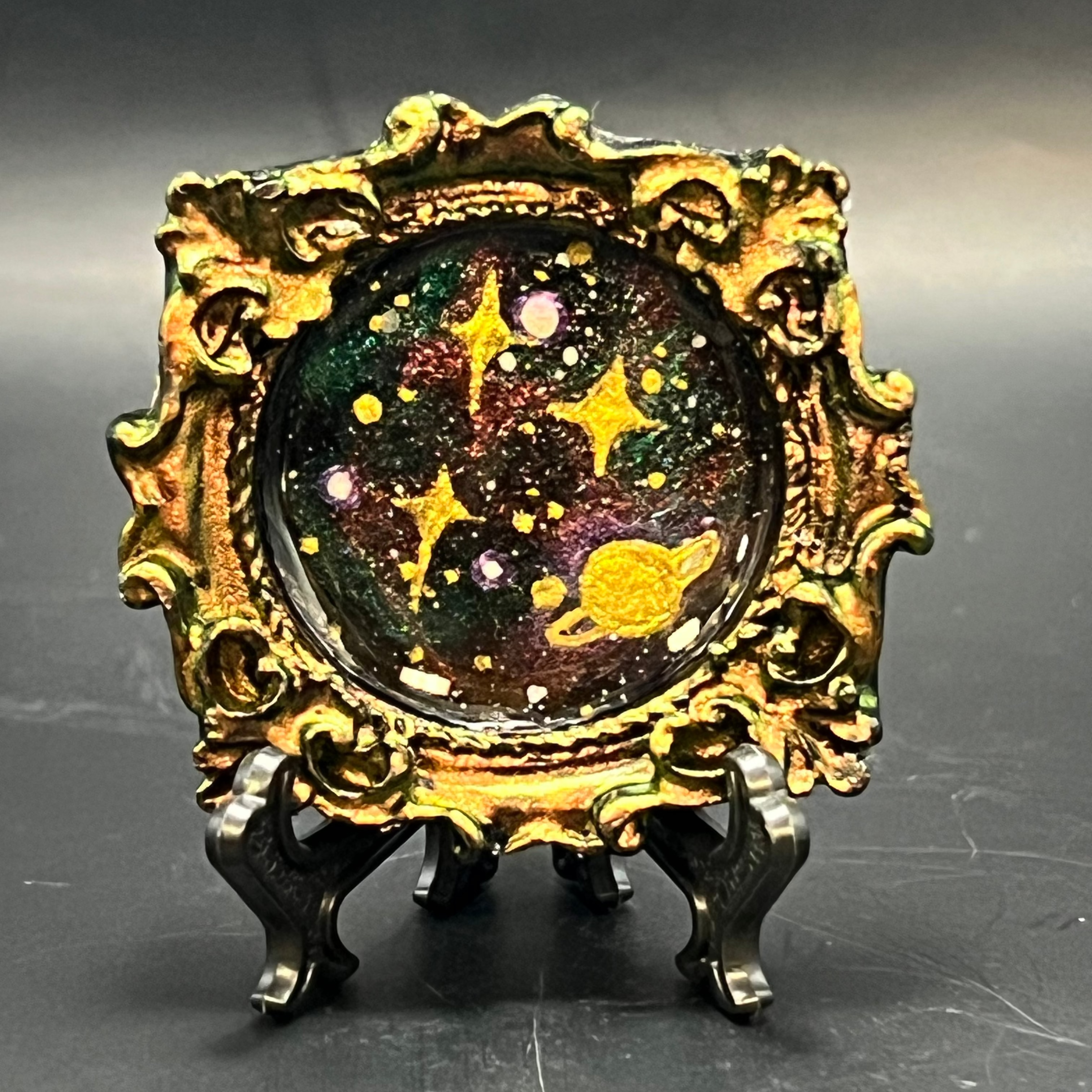 This is the handmade Ascension Miniature Painting by Chris Foster Design. This is showcasing the front. This is a decorative piece with a gold oval frame, starry night design including yellow stars, sparkling green, red, black and white smaller stars on a dark background. There is a magnet and against a miniature picture frame.