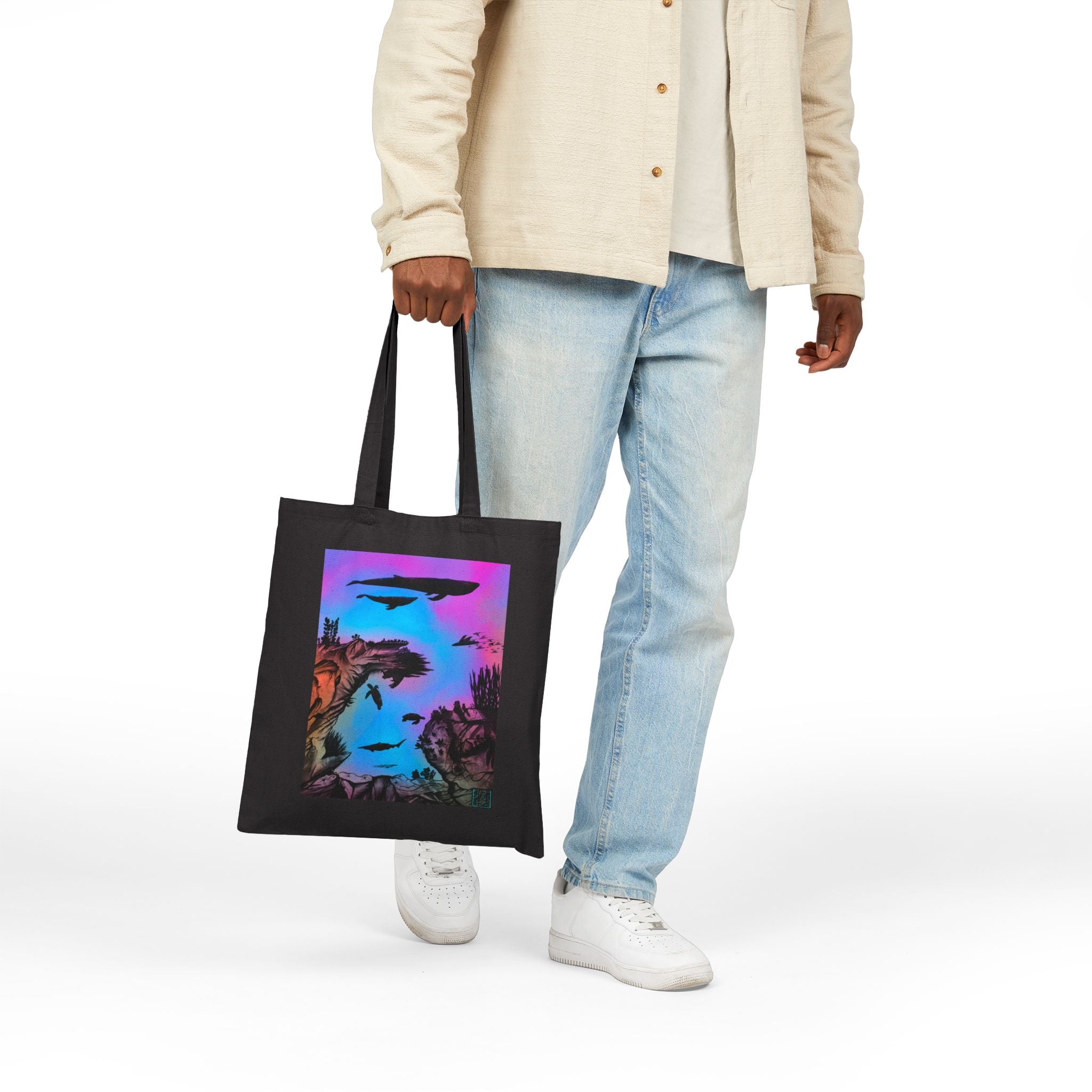 This is the Black Into the Abyss Canvas Tote by Lee Hansheng Studios.The illustration is underwater seascape with aquatic plants on a marbled background.There are two blue whales,1 goblin shark,school of eagle rays,two sea turtles and a school of fish.On the bottom right corner where the front patch of the backpack there is the studio logo printed in black outlined in blue.The colors are pink,purple,blue and red/orange.A guy is holding the tote in his left hand wearing white walking to the left.