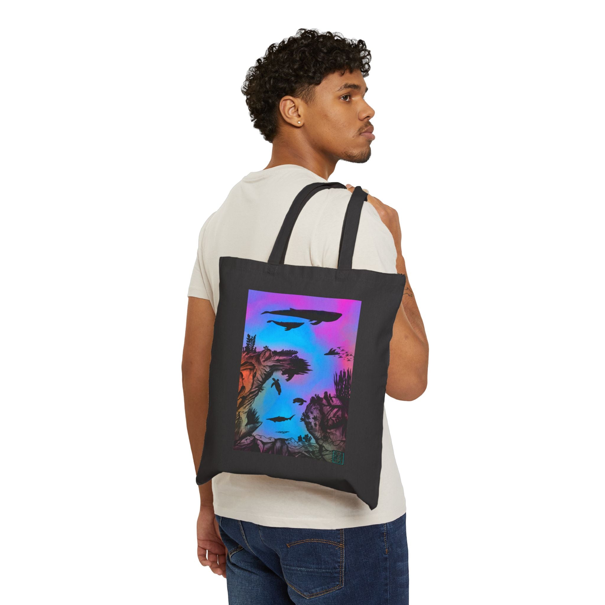 This is the Black Into the Abyss Canvas Tote by Lee Hansheng Studios.The illustration is underwater seascape with aquatic plants on a marbled background.There are two blue whales,1 goblin shark,school of eagle rays,two sea turtles and a school of fish.On the bottom right corner where the front patch of the backpack there is the studio logo printed in black outlined in blue.The colors are pink,purple,blue and red/orange.A man has the tote over his shoulder wearing jeans and white tee looking to the right.