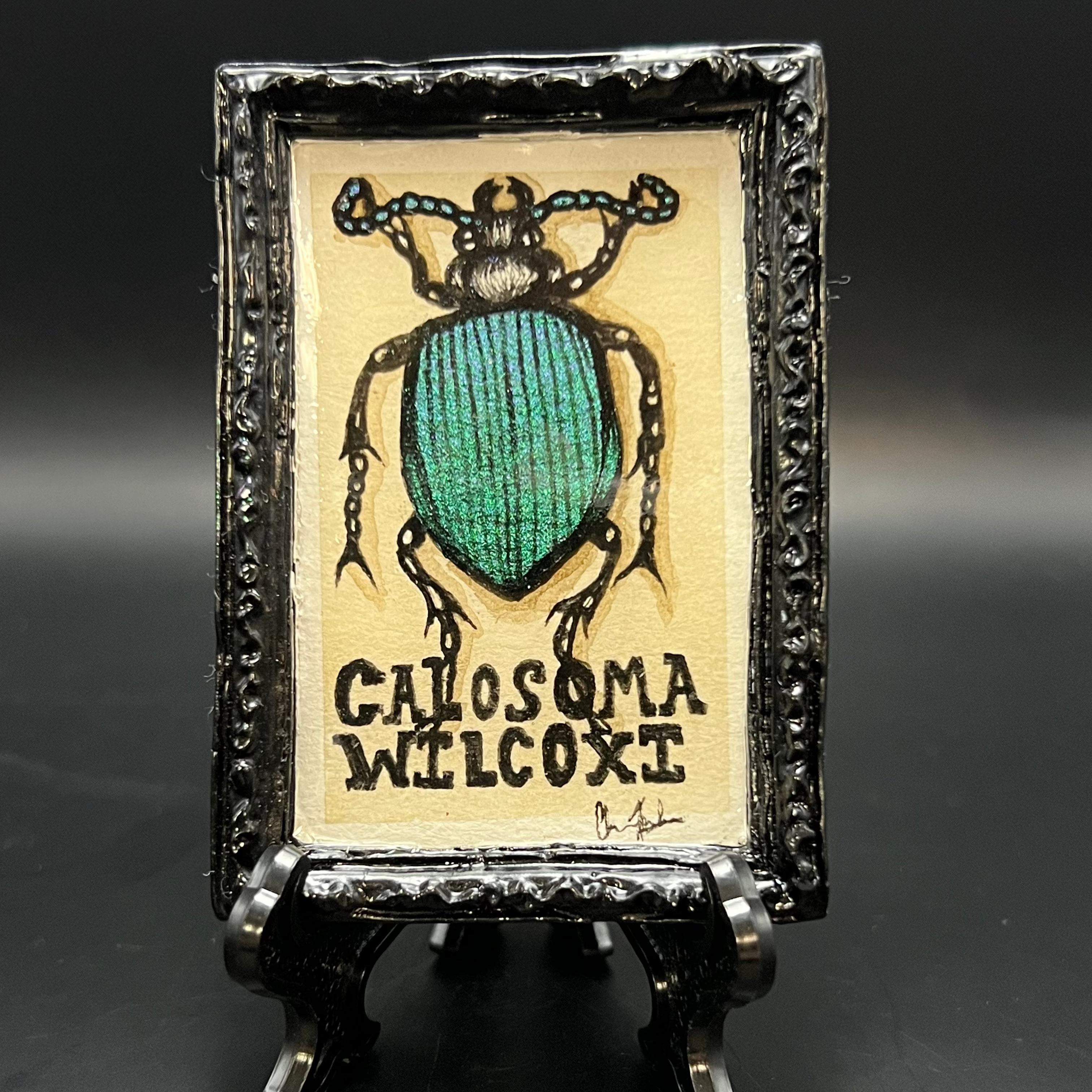 This is the Calasoma Wilcoxi Beetle Mini Painting by Chris Foster Design. This is showcasing the fron. High gloss black large rectangular mini painting. The Calosoma Wilcoxi beetle in all its glory painted in blue, purple, and green color shift watercolors to simulate its appearance in nature on a beige background with shadow accents. It is against a dark background.