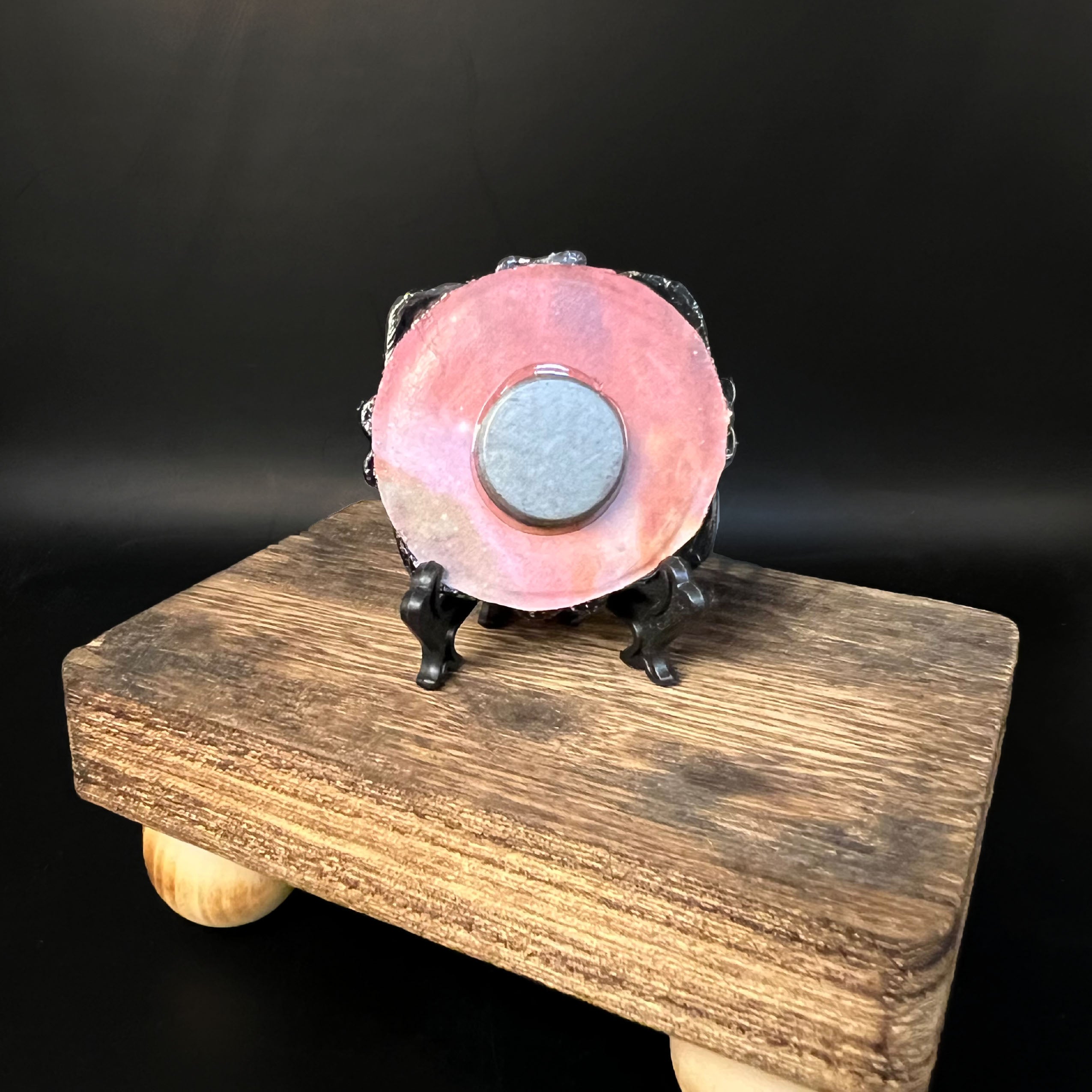 This is the Cat on Moon Mini Painting by Chris Foster Design. This is showcasing the back of the painting. The painting has a black circular frame backed with an orange,pink background with a round circular magnet and a black stand. It is on a wooden stand against a black background.