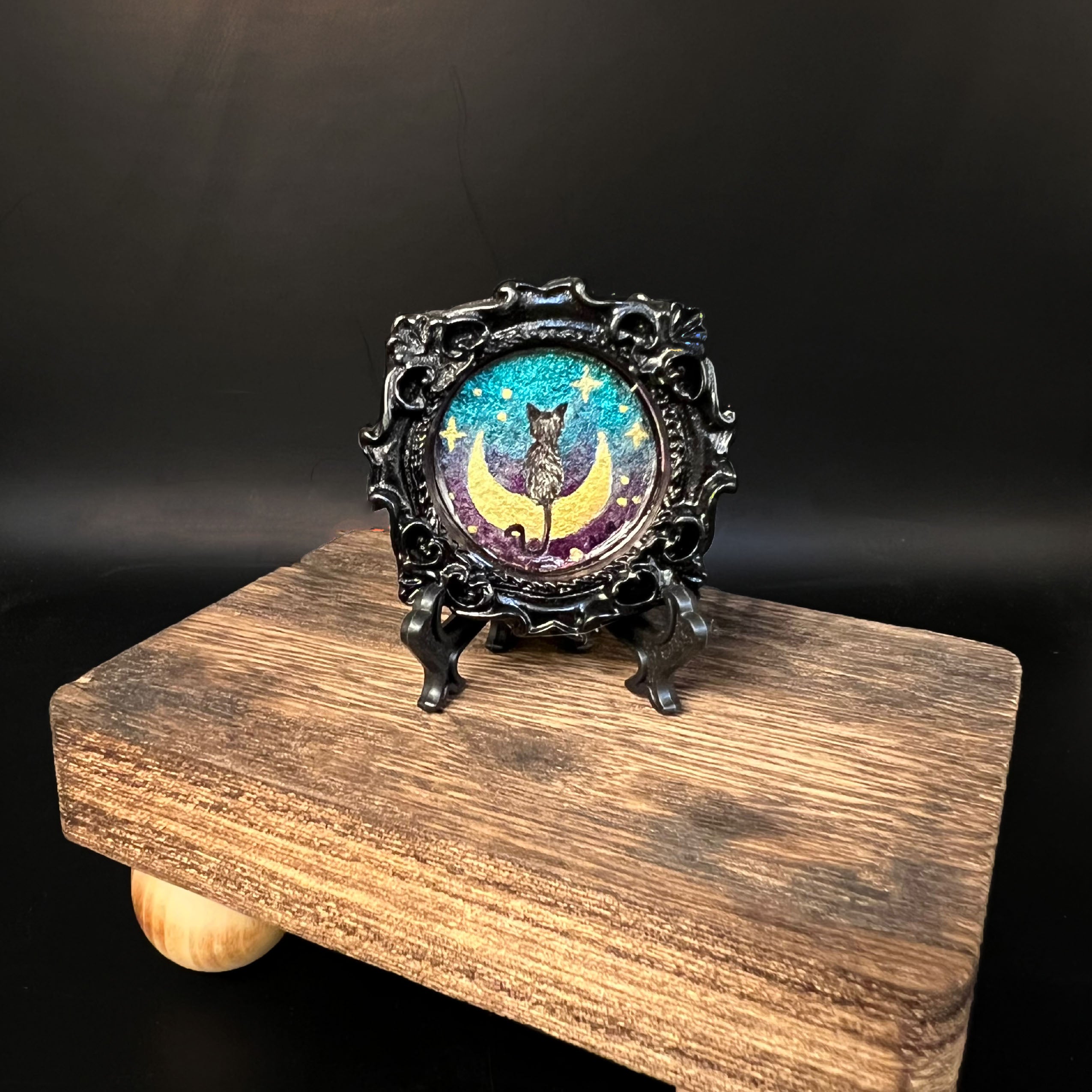This is the Cat on Moon Mini Painting by Chris Foster Design. This is showcasing the front of the painting. The painting has a black circular frame with a black cat sitting on a crescent moon, shimmery blue and purple background with yellow stars and dots. The painting is on a black stand. The miniature painting in general is on a wooden stand against a dark background.