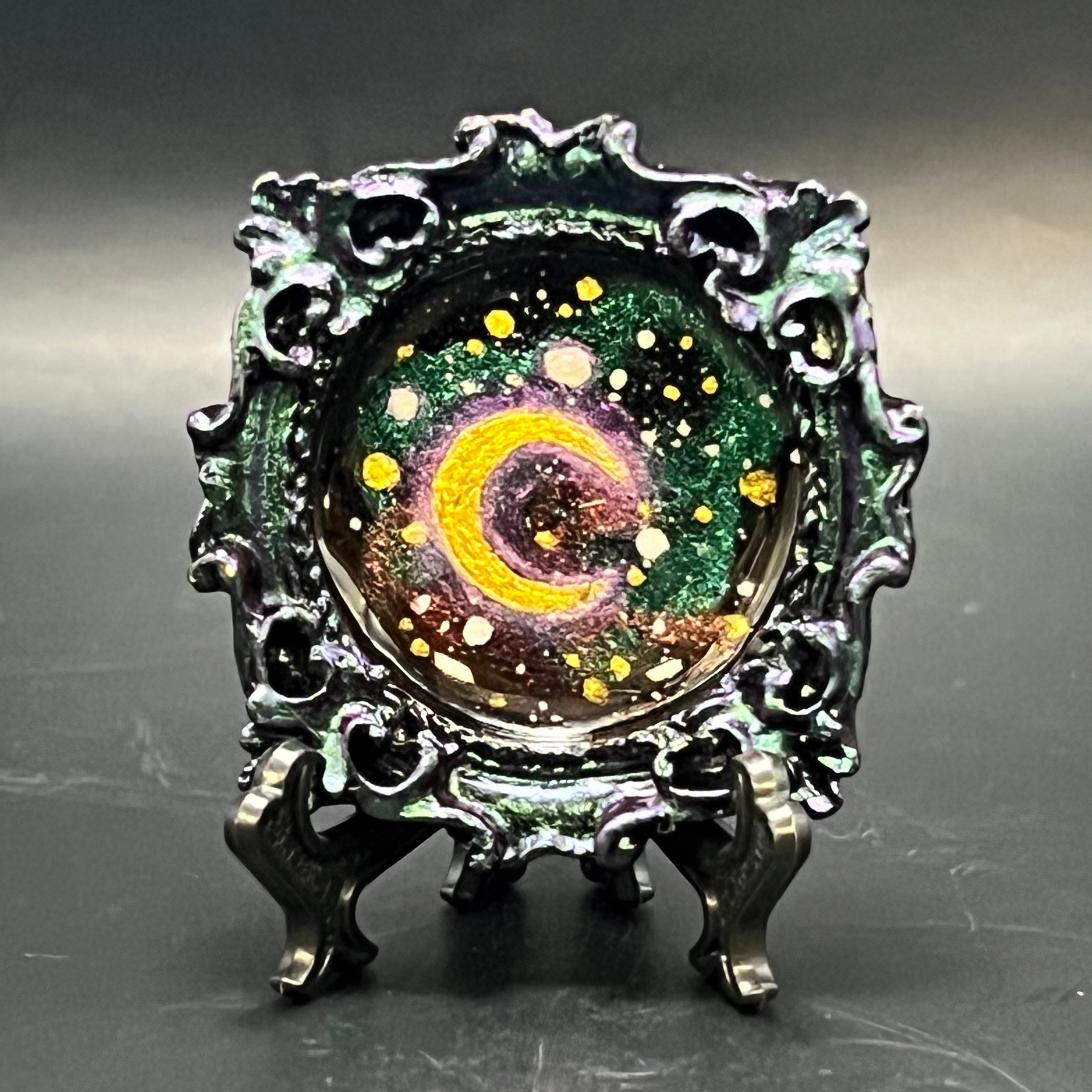 This is the Clustered Crescent Mini Painting by Chris Foster Design. This is showcasing the front. Pink gold encrusted black circle frame. A glowing golden crescent moon floats with friends amongst silver gold and white stars upon pink gold and purple blue color shifting nebulae. There's a black stand and is against a dark background.