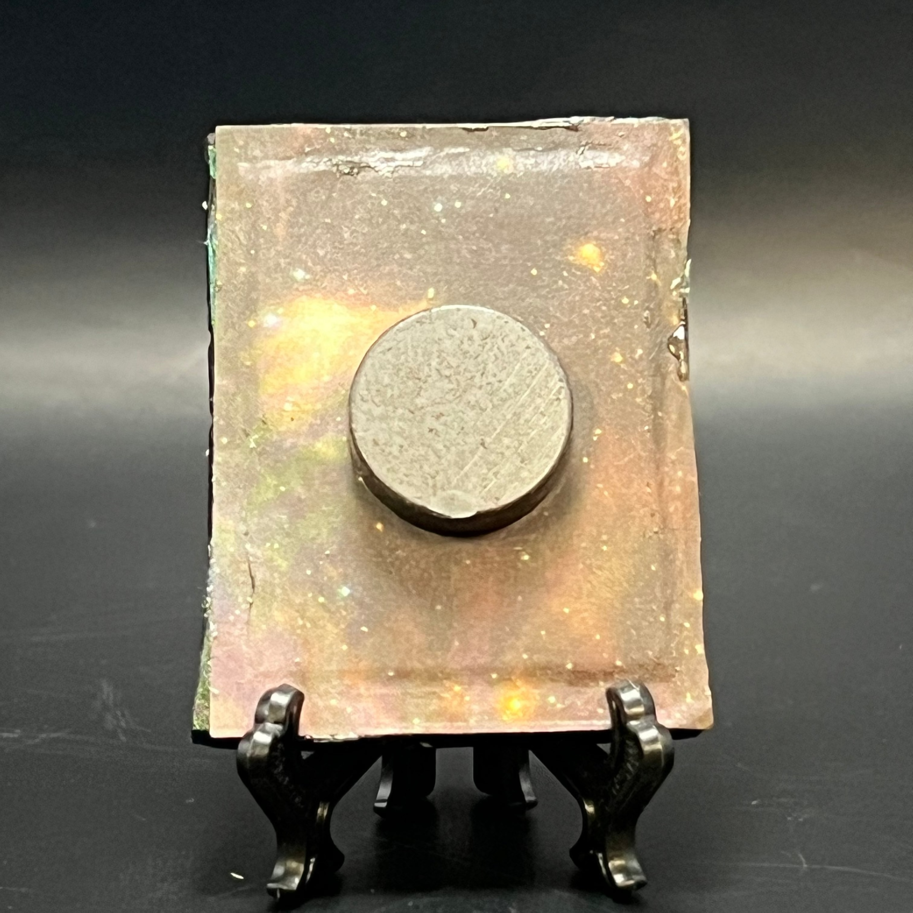 This is the handmade Distant Ringed Planet Miniature Painting by Chris Foster Design. This is showcasing the back. This is a small rectangular frame and the decorative item with a sparkly mix background with orange, yellow and black on a stand with yellow stars. There is a magnet and against a miniature picture frame.