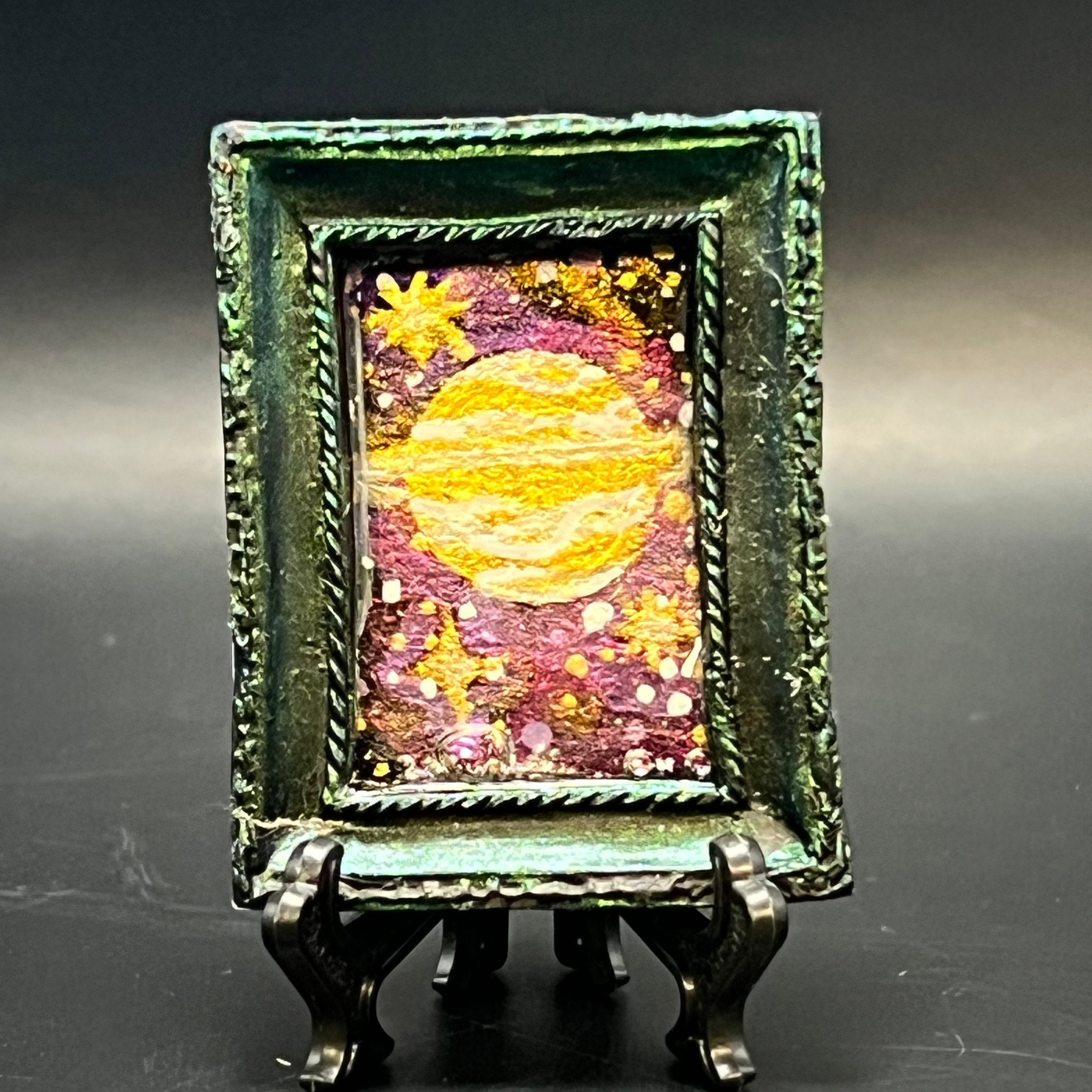 This is the handmade Distant Ringed Planet Miniature Painting by Chris Foster Design. This is showcasing the front. This is a decorative piece with a shimmery green rectangular frame, starry night design including yellow stars, sparkling red, orange, yellow and white stars on a dark background. There is a magnet and against a miniature picture frame.