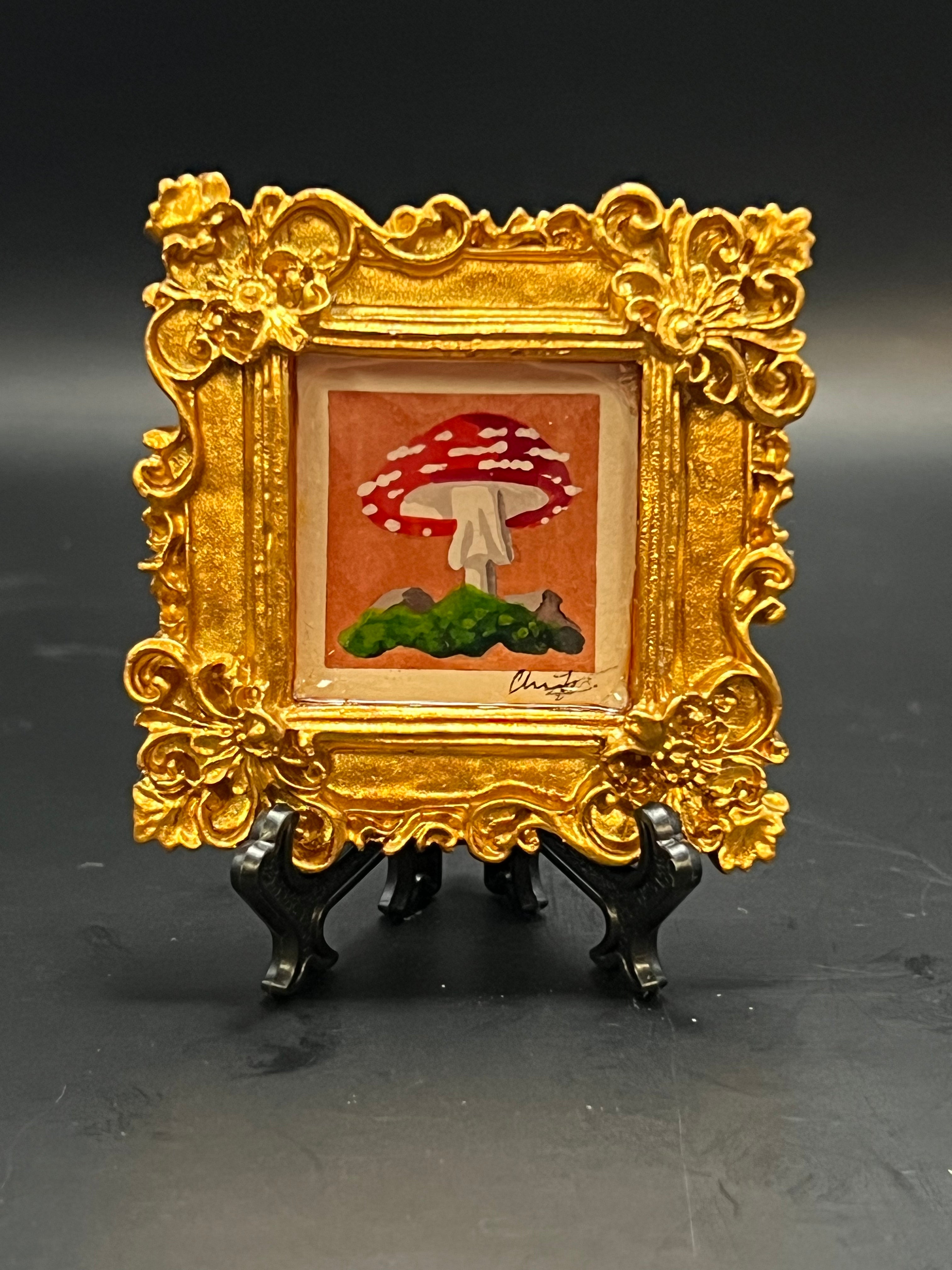 This is the Fly Agaric Mushroom Orange Miniature Painting by Chris Foster Design. This is a small painting of one mushroom that is red and speckled white, dark orange background and wavy dark green grass in a decorative gold square frame on a black stand.