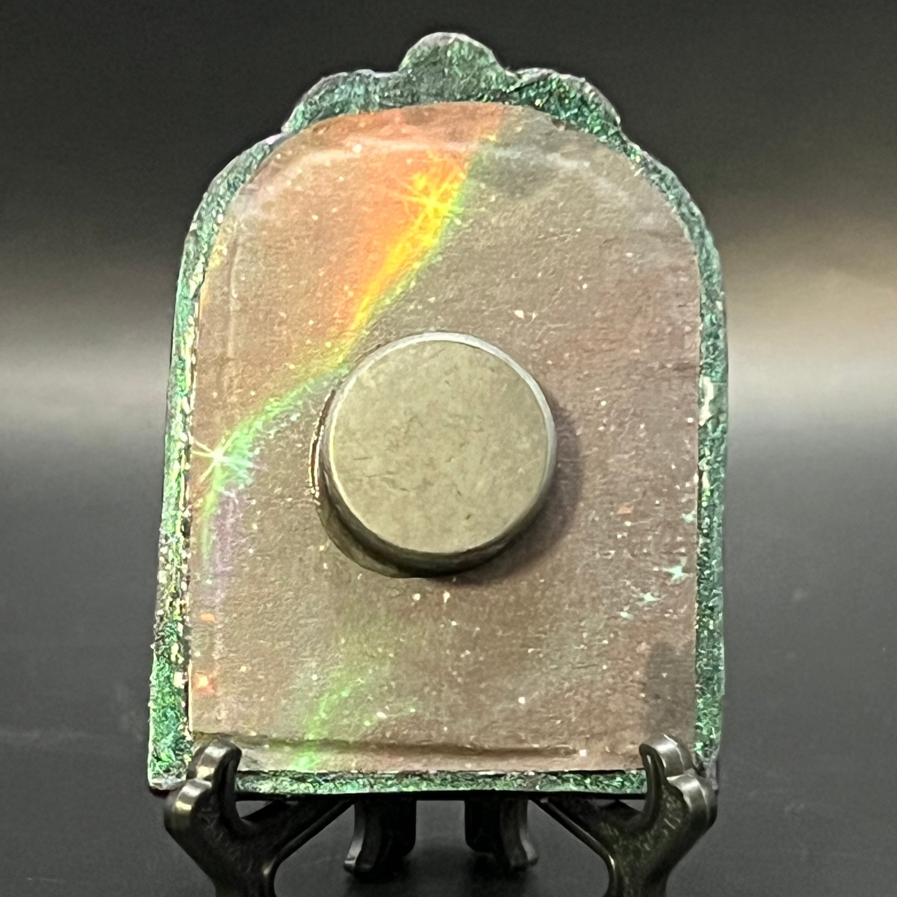 This is the handmade Gem in the Void Miniature Painting by Chris Foster Design. This is showcasing the back. Decorative piece with a metallic base on a dark background on a stand. There is a magnet and against a miniature picture frame.