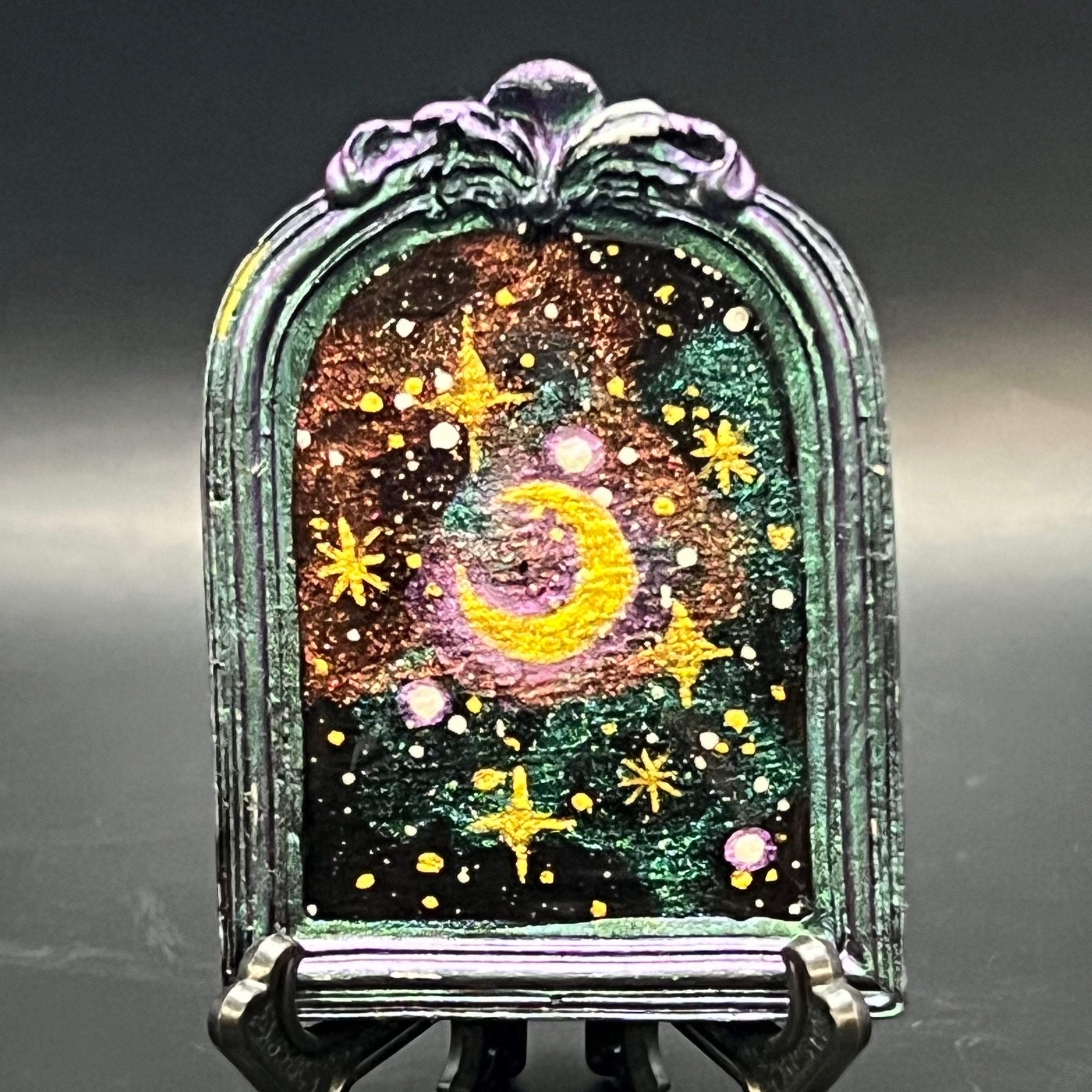 This is the handmade Gem in the Void Miniature Painting by Chris Foster Design.This is showcasing the front. It is a decorative piece with a celestial design featuring stars and a moon on a stand against a dark background. The colors are sparkling yellow, orange, green and black that swirl around. There is a magnet and against a miniature picture frame.