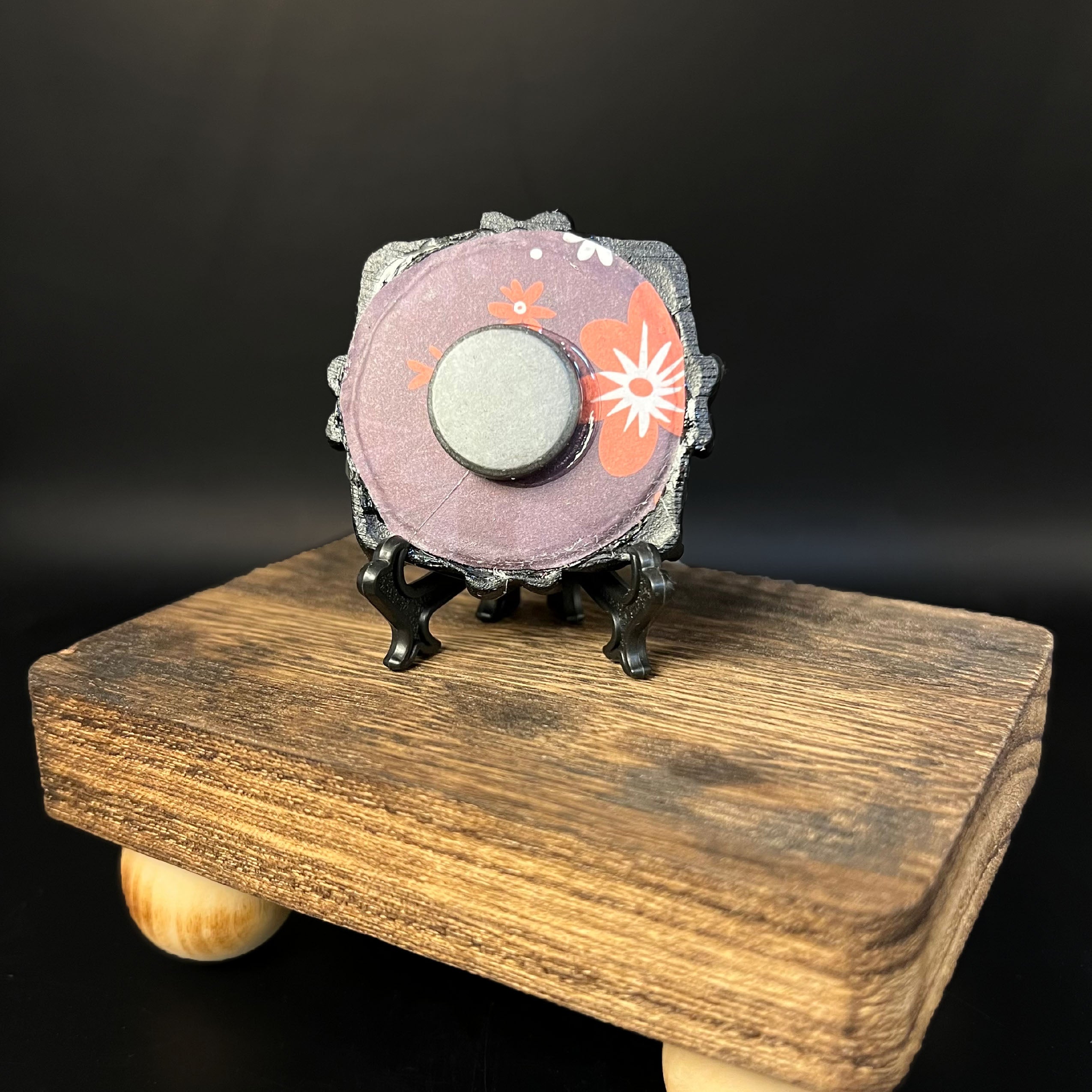 This is the Glistening Web Mini Painting by Chris Foster Design. This is showcasing the back of the painting. The painting has a black circular frame backed with a maroon and flowery background. The painting is on a black stand and has a round magnet in the middle. The miniature painting in general is on a wooden stand against a dark background.