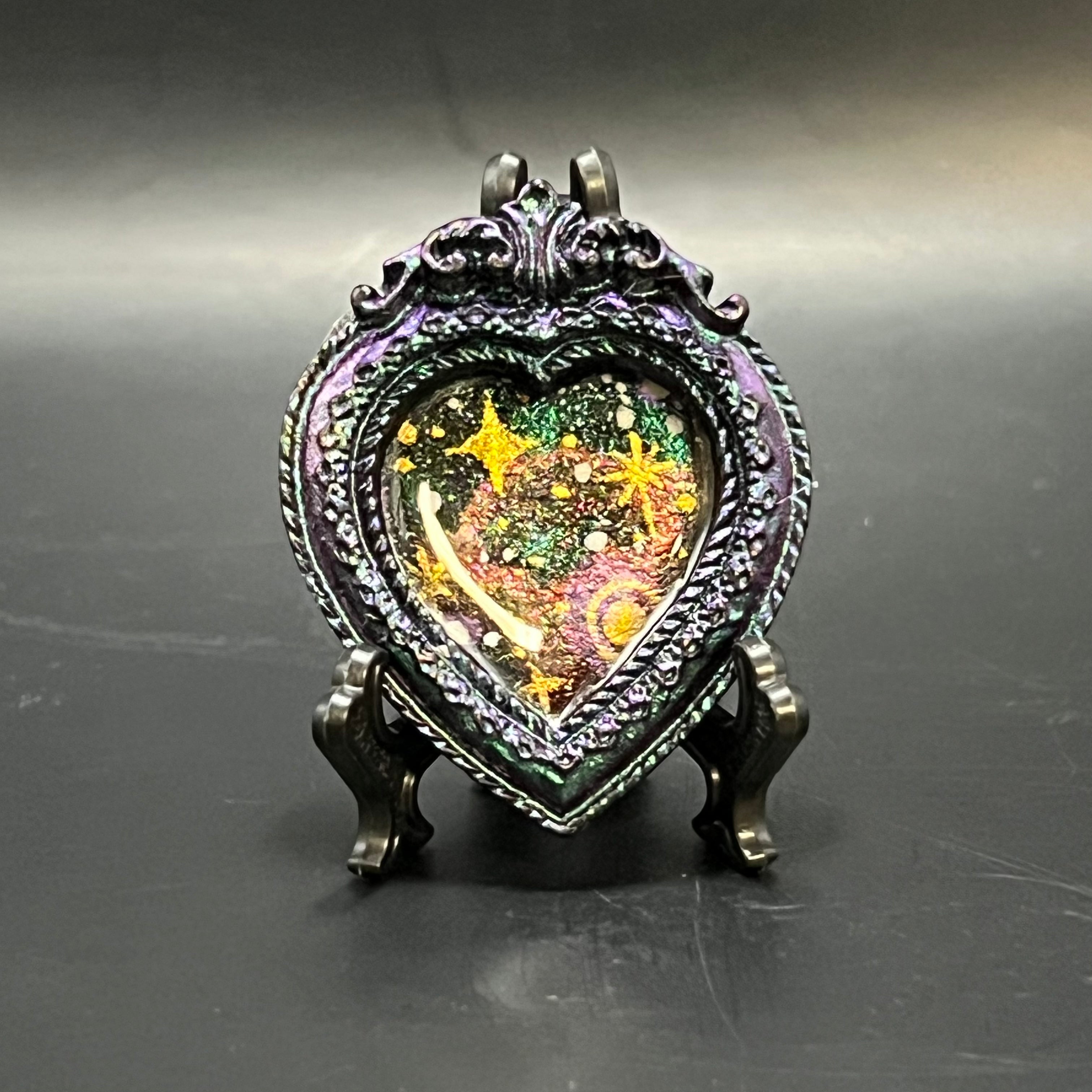 This is the Glowing Cluster Mini Painting by Chris Foster Design. This is showcasing the front. The decorative heart-shaped frame Glowing cluscter handmade painting by Chris Foster Design. Purple heart shaped frame with an illustration of brilliance of colors and stars sealed with resin. It is on a black stand and against a dark background.