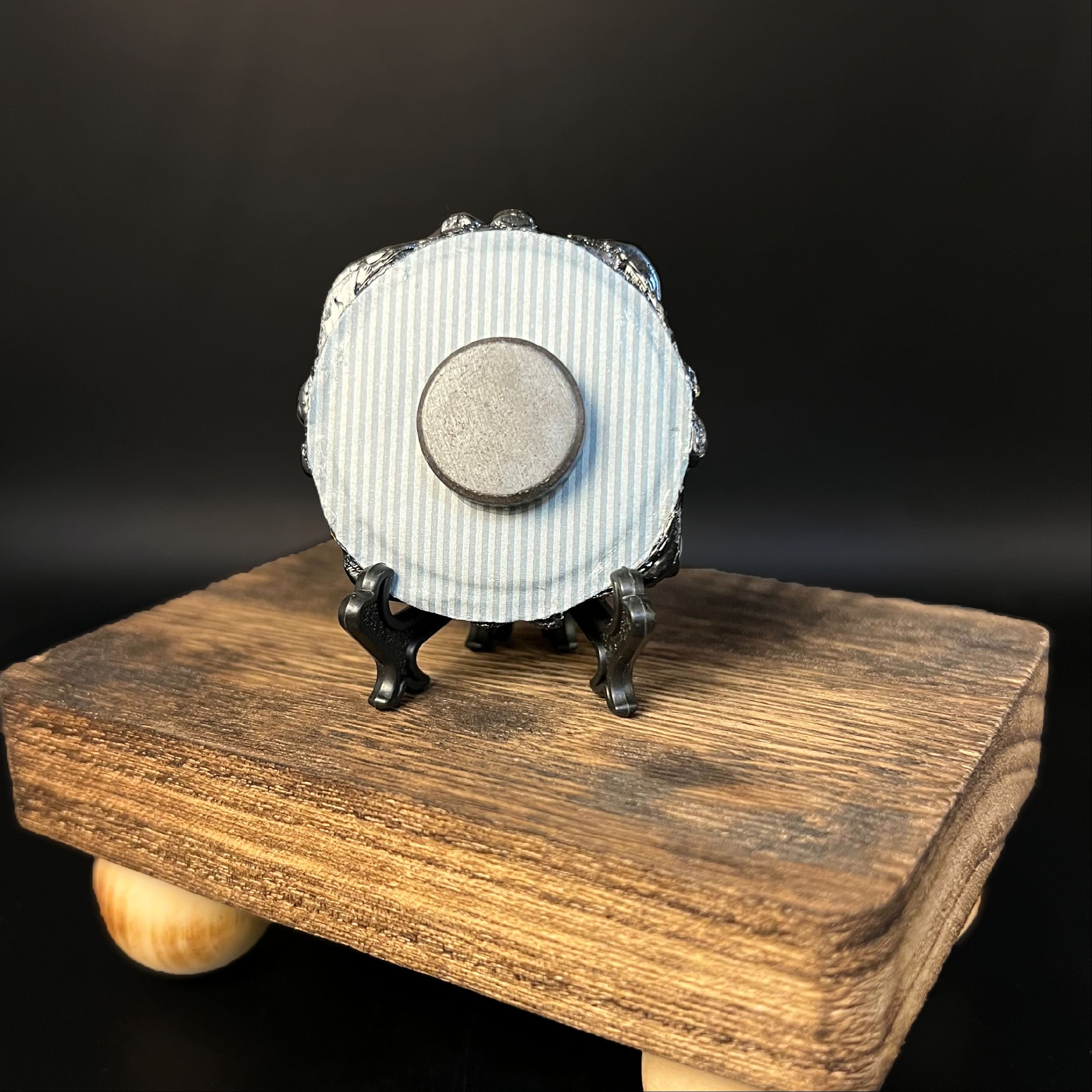 This is the Green Bat Mini Painting by Chris Foster Design. This is showcasing the back of the painting. The painting has a black circular frame backed with a blue and white striped background. A round magnet in the middle and painting overall on a black stand. It is on a wooden stand board against a dark background.