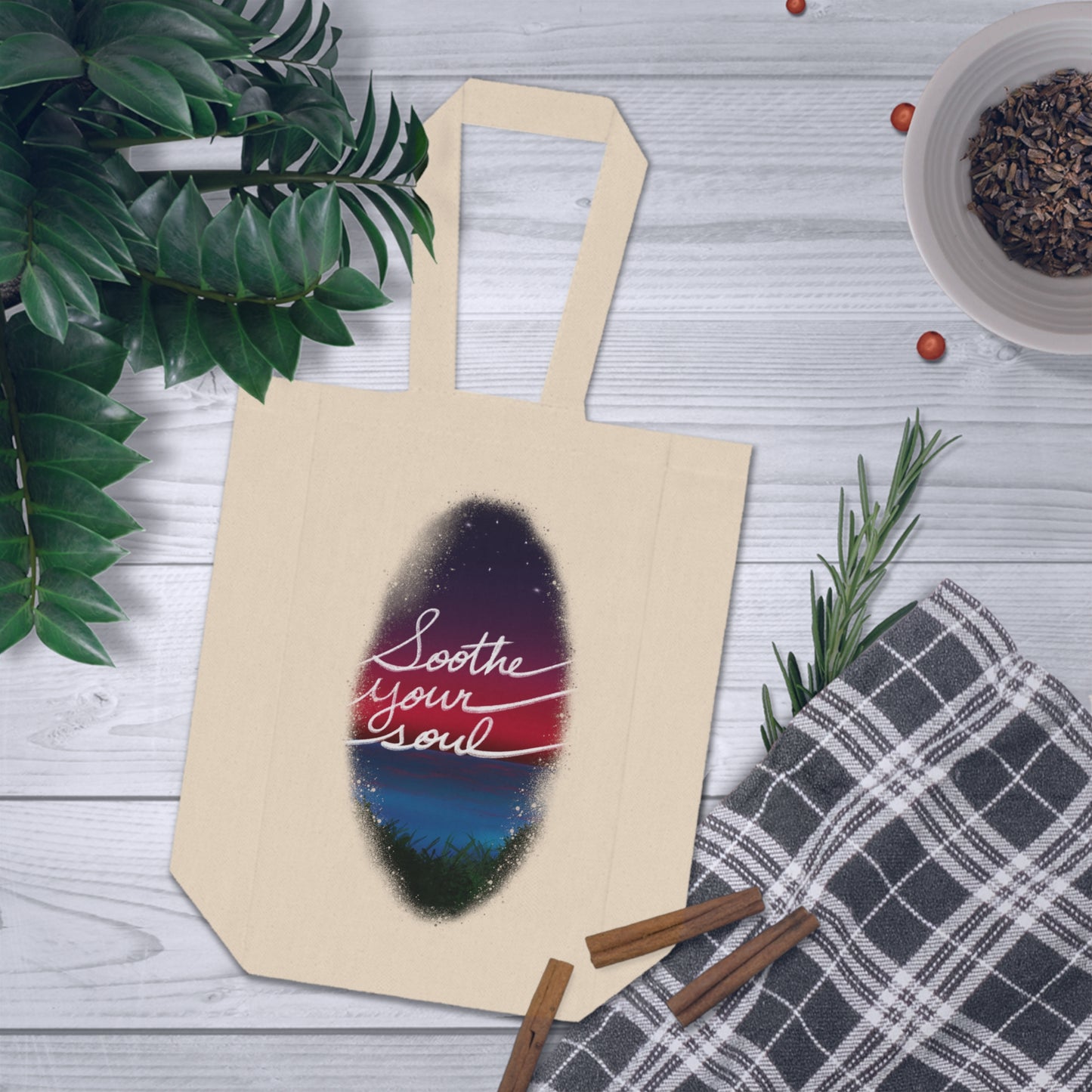 The Natural one size Soothe Your Soul Double Wine Tote Bag by Lee Hansheng Studios.This tote securely holds two wine bottles and image showcases the tote from the front.The illustration shows a faded oval drawing of the night sky with white twinkling stars, pink,purple and blue colors.The words shows,"soothe your soul".The tote is on a white wood table,left has luscious dark green plants,a bowl of shredded chocolate on the right,below that is a plaid grey and white blanket with three cinnamon sticks.