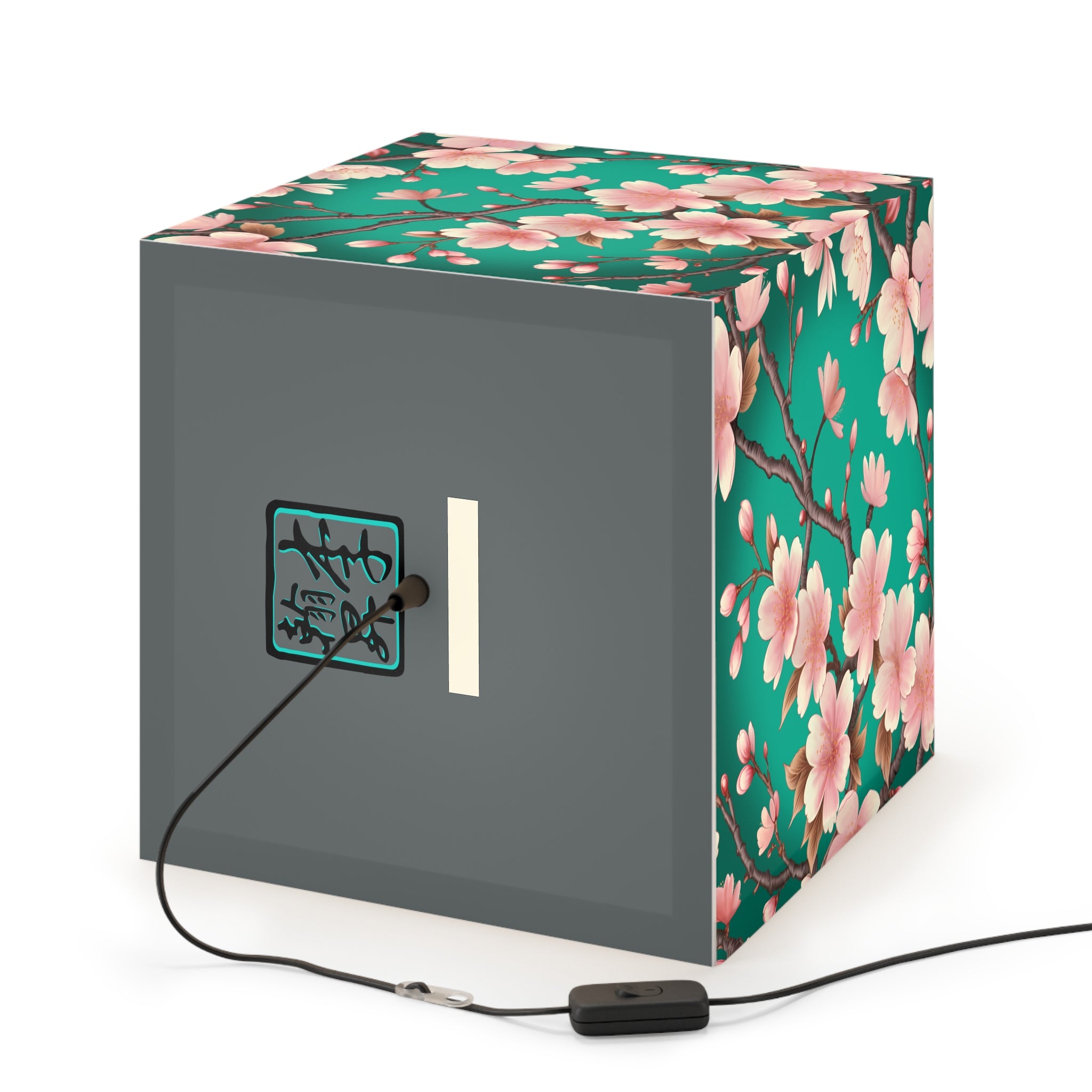 This is the Pendant Sakura Light Cube Lamp by Lee Hansheng Studios. The illustration is all around has a blue/teal background, 3-4 thin dark wood branches with pink/light pink buds on each stem. There are blooming and already flowered sakuras on each branch. There is about 3-4 inches between the end of the illustration and the sides of the lamp. The cord has a sleeker and plug-free option. The cube lamp is on a gray back where the logo and cord is plugged in. It is against a white background.