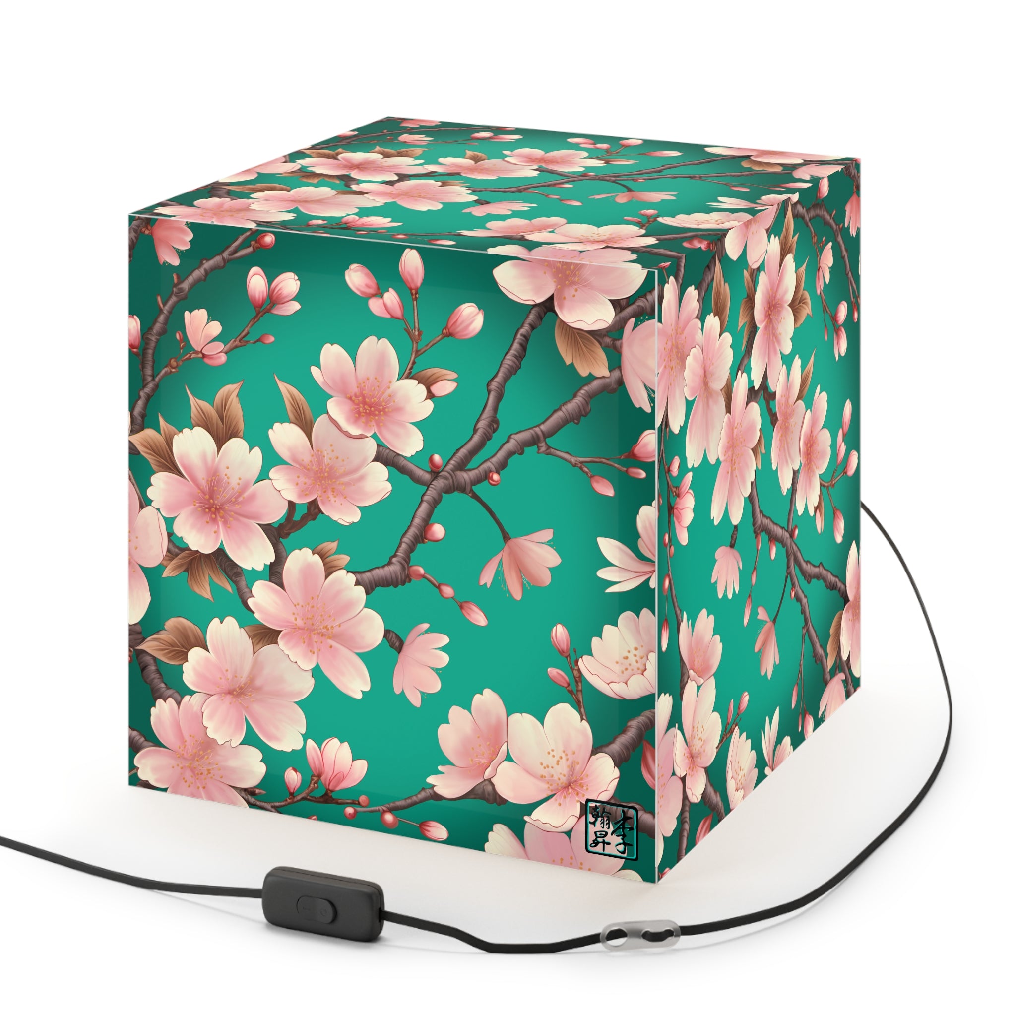 This is the Pendant Sakura Light Cube Lamp by Lee Hansheng Studios. The illustration is all around has a blue/teal background, 3-4 thin dark wood branches with pink/light pink buds on each stem. There are blooming and already flowered sakuras on each branch. There is about 3-4 inches between the end of the illustration and the sides of the lamp. The cord has a sleeker and plug-free option. The cube lamp's top is facing us. It is against a white background.