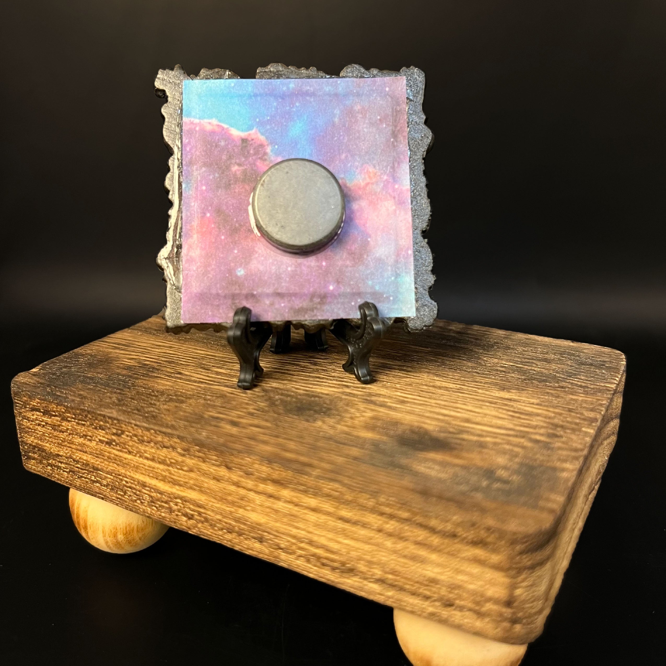 This is the Pentacle Mini Painting by Chris Foster Design. This is showcasing the back of the painting. It is backed by a galaxy design in blue, red, orange and dark blue with a round magnet in the middle. The miniature painting is resting on a black stand and the whole is on a wooden stand against a dark background.