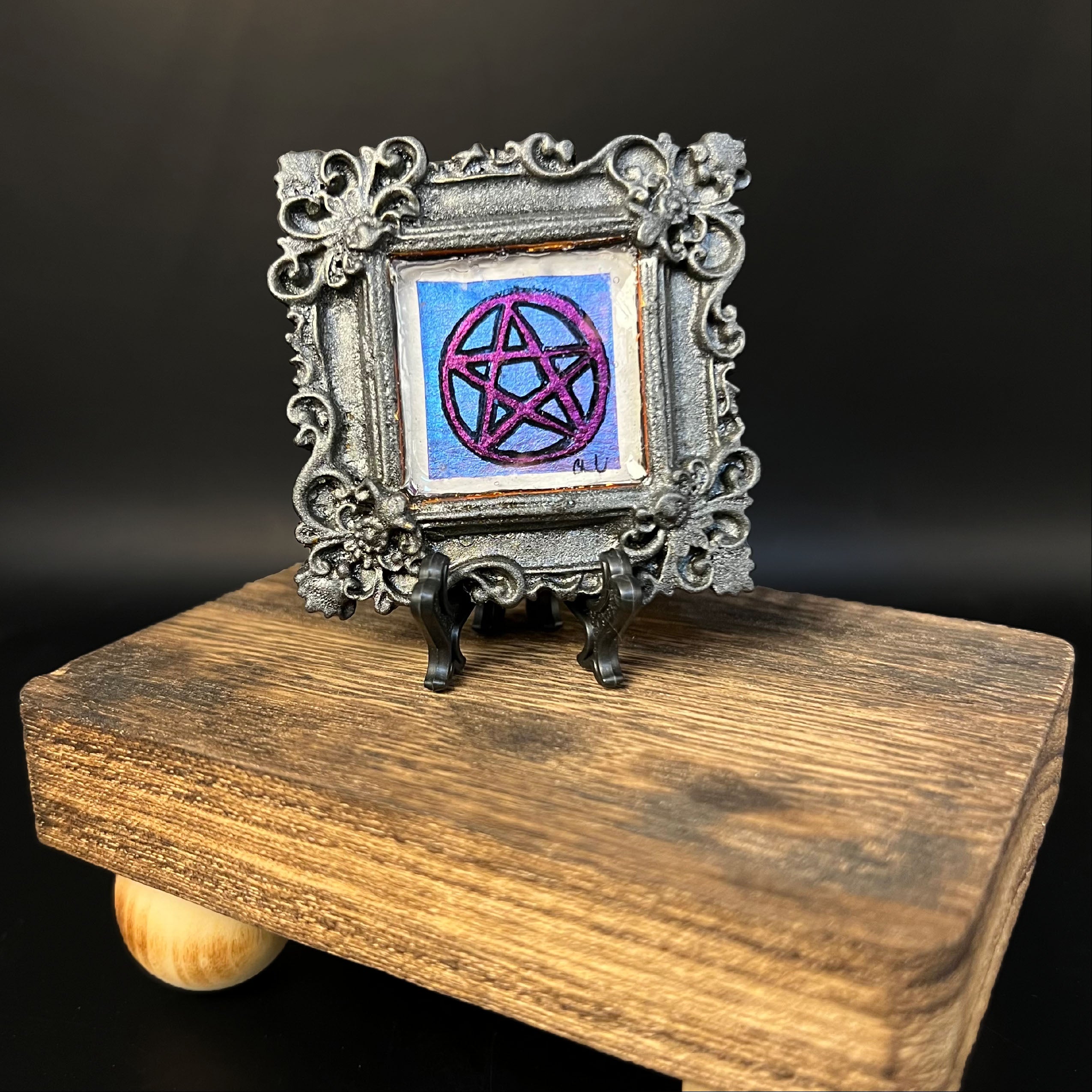This is the Pentacle Mini Painting by Chris Foster Design. This is showcasing the front of the painting. There is a rectangular muted black with sparkles frame with a purple and black pentagram design. The background of the pentagram is blue with the studio's signature on the bottom right. It is on a wooden stand against a dark background.