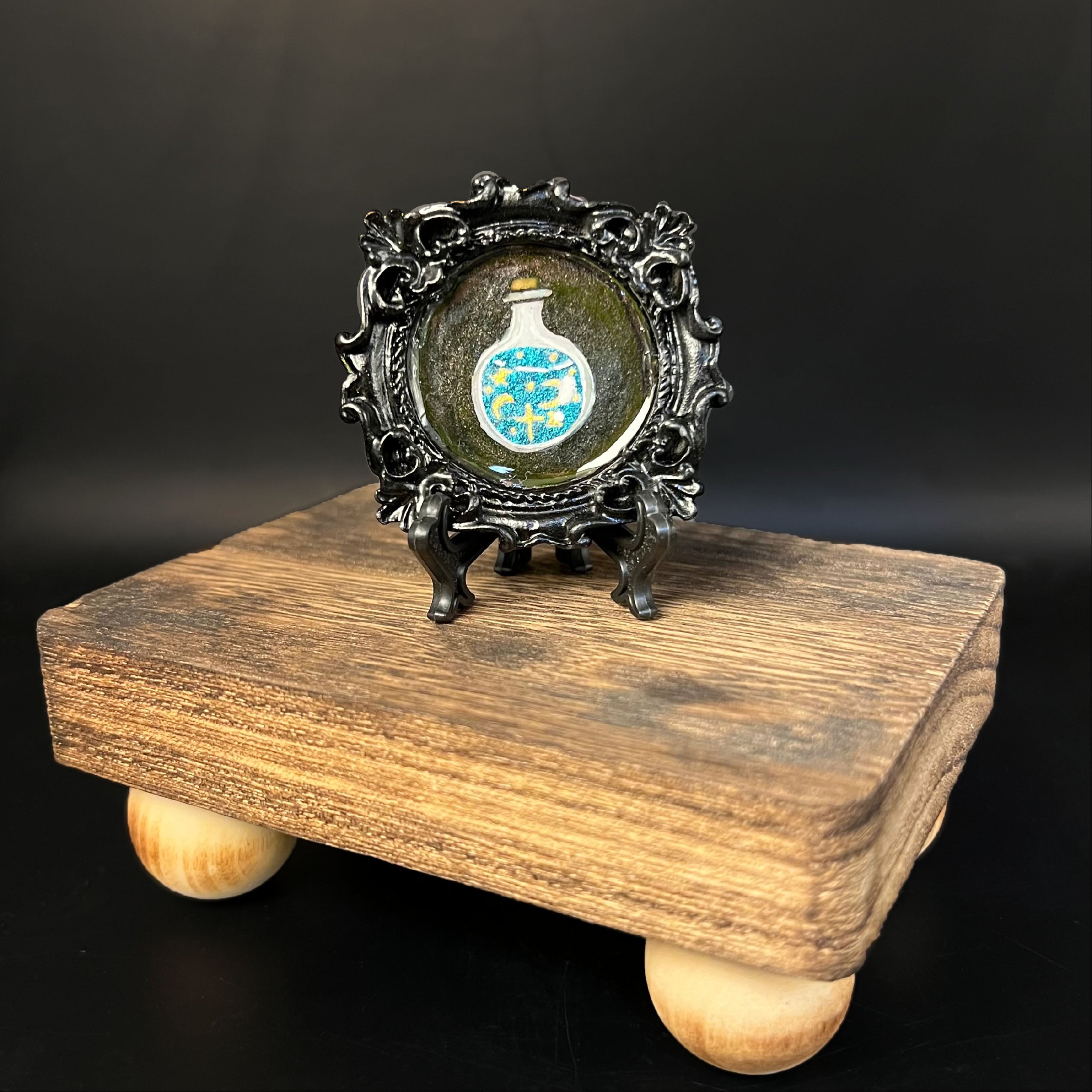 This is the Potion Mini Painting by Chris Foster Design. This is showcasing the front of the painting. The painting has a black circular frame with a potion bottle that is clear and a tan cork. The bottle is filled with shimmery blue liquid and yellow moon, stars and dots. The painting is on a black stand. The miniature painting in general is on a wooden stand against a dark background.