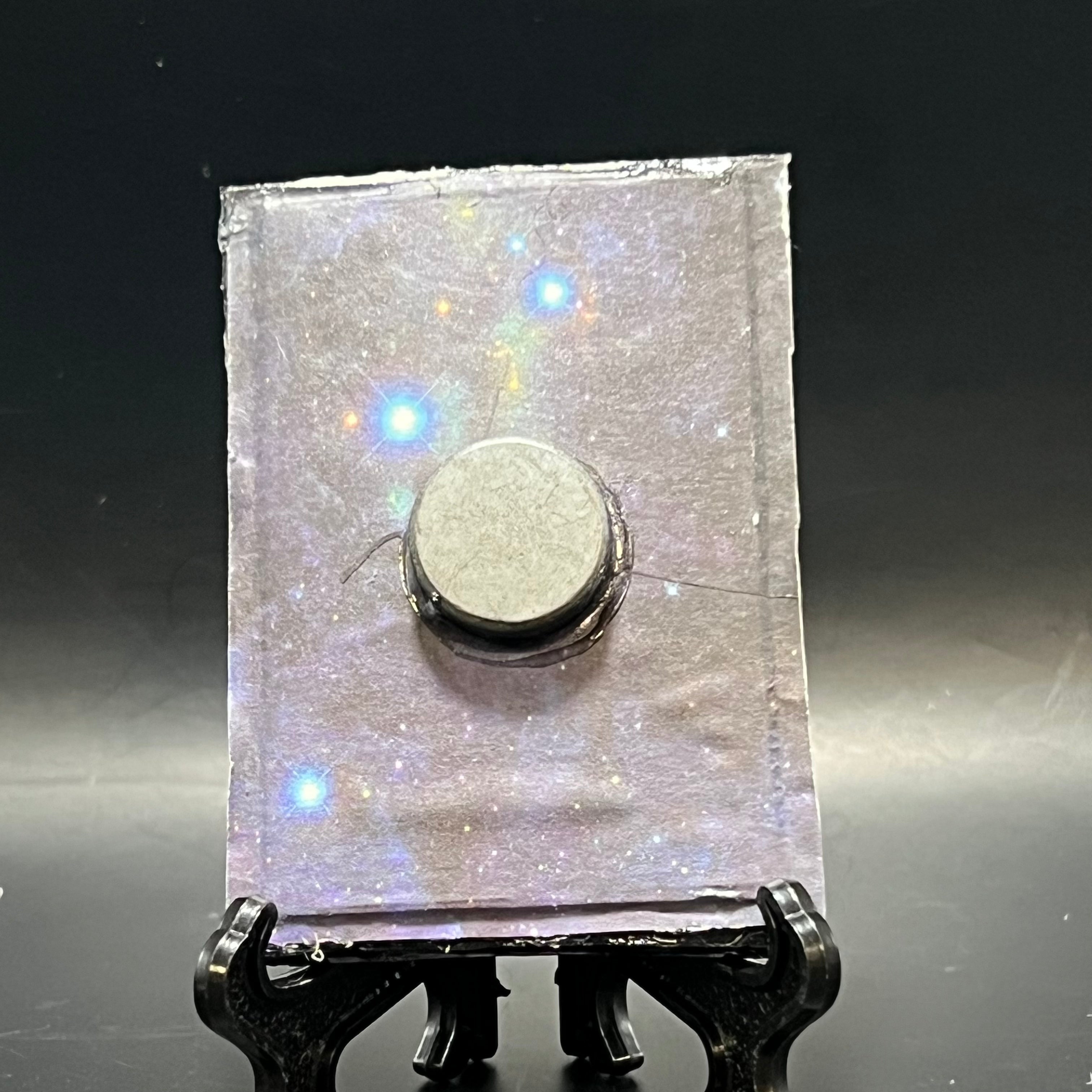 This is the Saturn in Retrograde Mini Painting by Chris Foster Design. This showcases the back. Backed with black and blue space void origami paper. It is against a dark background.