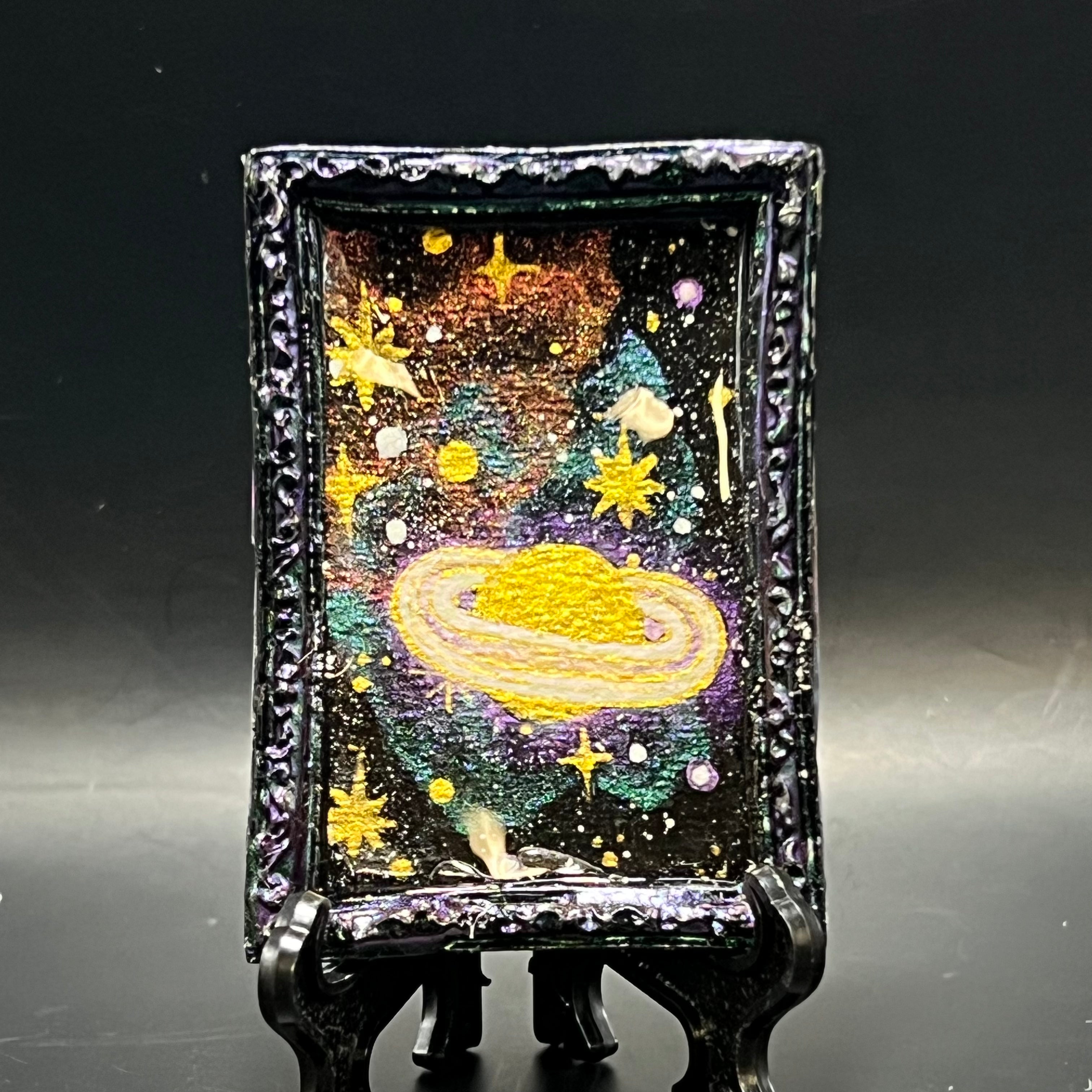 This is the Saturn in Retrograde Mini Painting by Chris Foster Design. This is showcasing the front. Black rectangular frame encased in red purple color shift paint. A gold and white saturn floats high in the void of space amongst white and glowing gold stars, set upon red purple and gold green, color shifting nebulae. It is against a dark background.