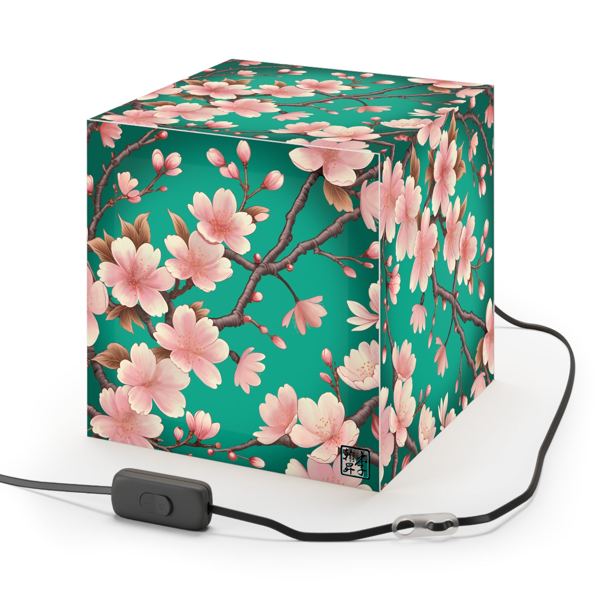 This is the Standard Sakura Light Cube Lamp by Lee Hansheng Studios. The illustration is all around has a blue/teal background, 3-4 thin dark wood branches with pink/light pink buds on each stem. There are blooming and already flowered sakuras on each branch. There is about 3-4 inches between the end of the illustration and the sides of the lamp. This includes plug for US or EU. The cube lamp is shown from the top. It is against a white background.