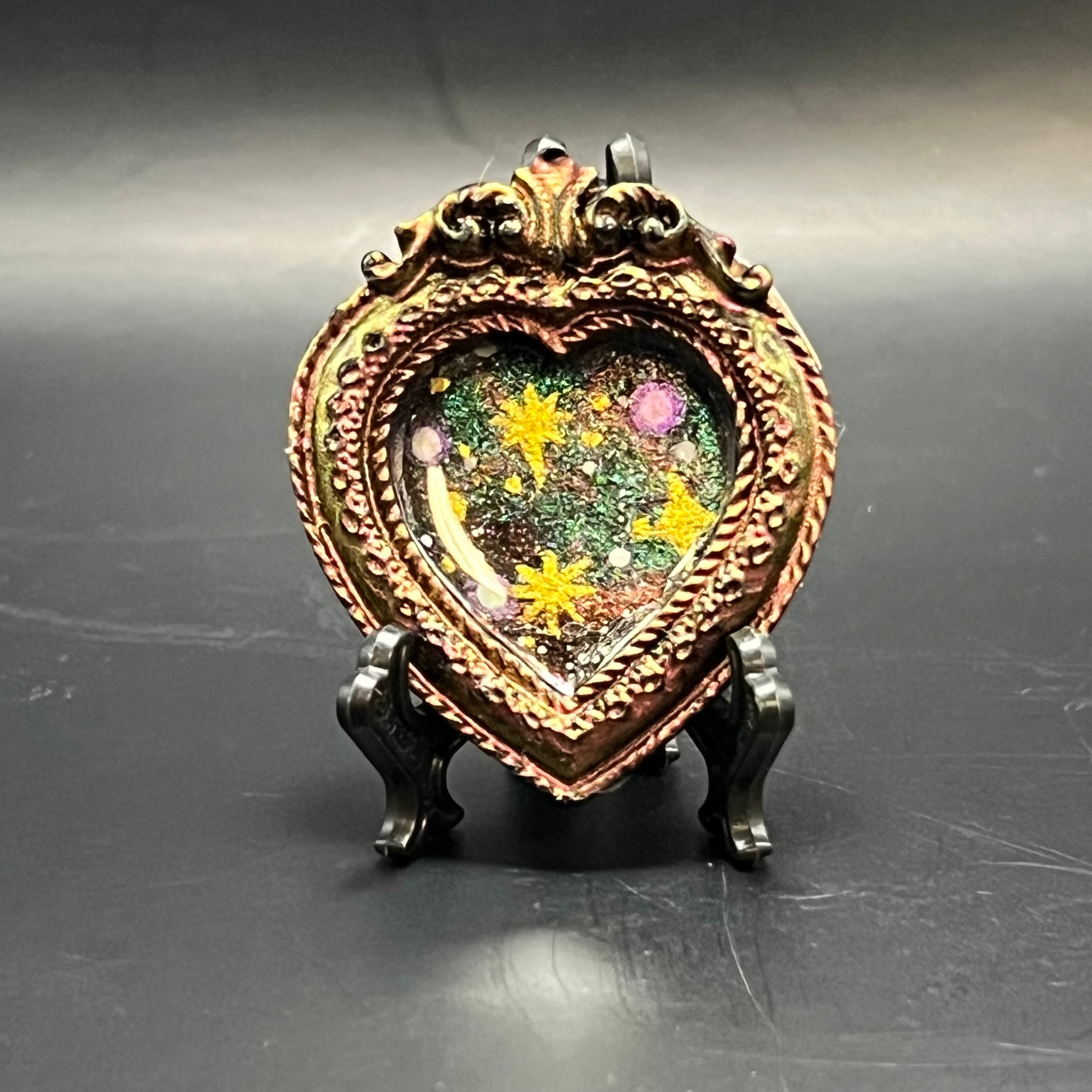 This is the Three Sisters Mini Painting by Chris Foster Design. This is showcasing the front. Brilliant gold pink and green color shift on this black heart frame filled with wonder from the void. Three glowing gold sister stars come together amongst a backdrop of white distant stars and color shifting pink gold purple and blue. It is against a dark background.