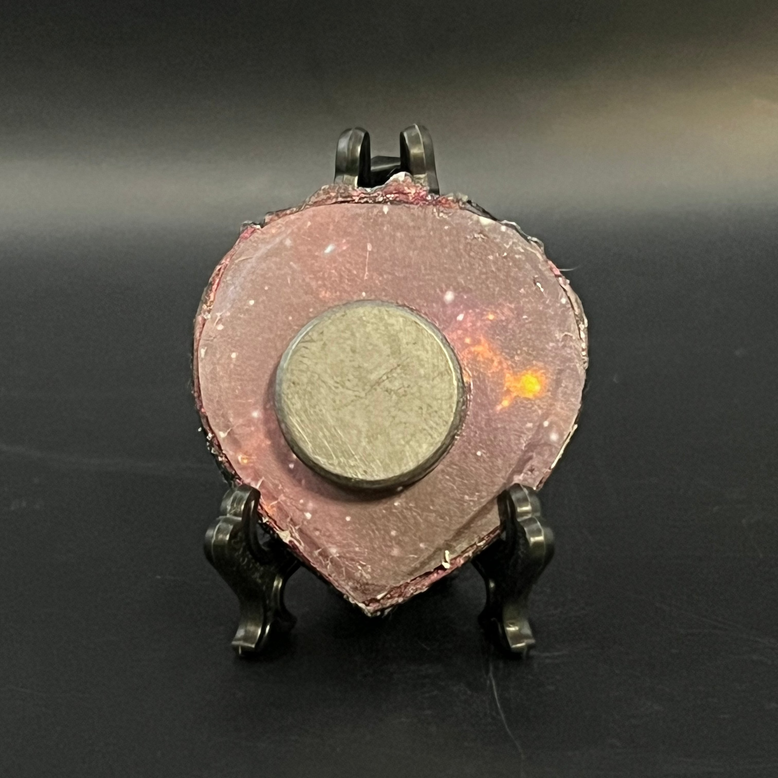 This is the handmade Twinkle in the Void Miniature Painting by Chris Foster Design. This is showcasing the back. This is a small heart frame and the decorative item with a sparkly mix background with black, orange and little stars on a stand. There is a magnet and against a miniature picture frame.