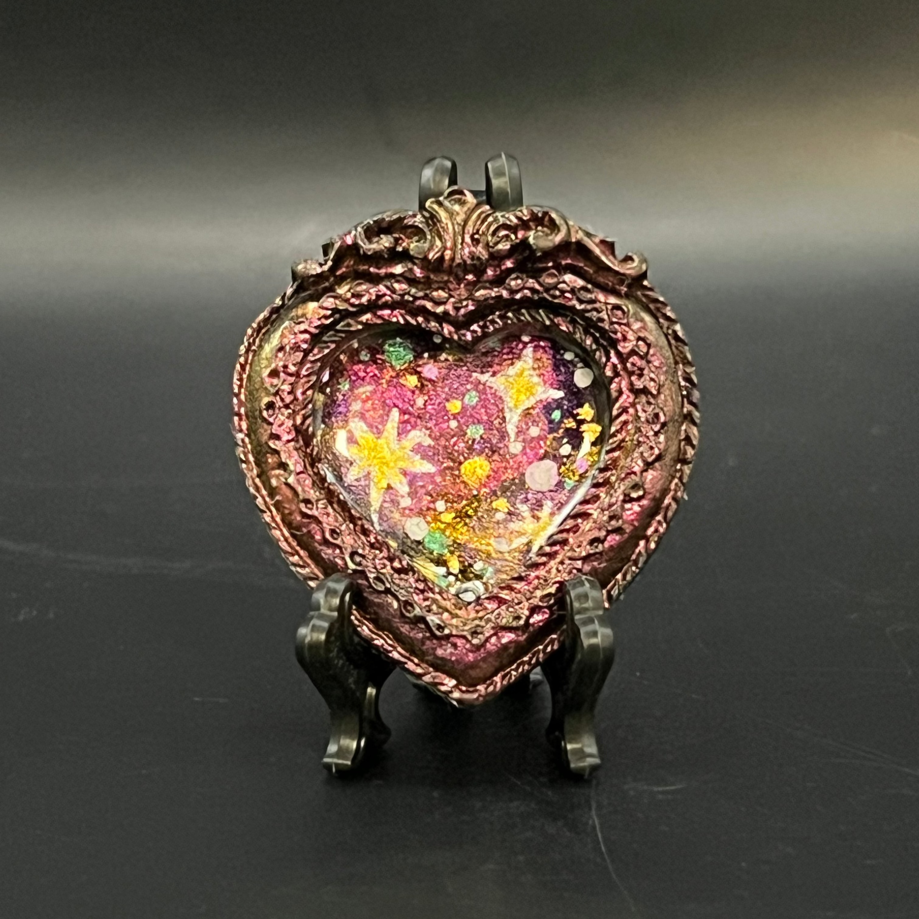 This is the handmade Twinkle in the Void Miniature Painting by Chris Foster Design. This is showcasing the front. This is a decorative piece with a shimmery red heart frame, starry night design including yellow stars, sparkling red mostly with, orange, black, green and white on a dark background. There is a magnet and against a miniature picture frame.