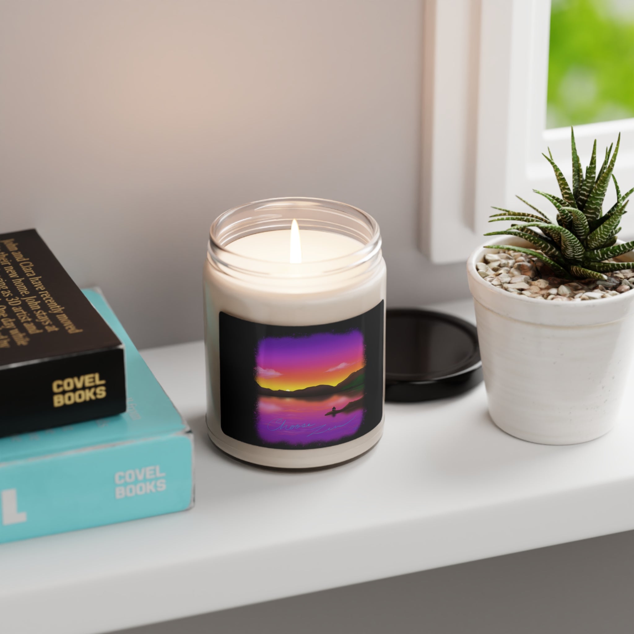 This is the Unscented Choose Zen Scented Soy Candle by Lee Hansheng Studios. The illustration is a landscape when the sun is setting from a lake.It's surrounded by black like an old time photo and phases into purple,pink,red,orange then yellow where you can see the darker green hills.The candle has a 'Choose Zen' illustration on the label in blue. The candle is on top of the white surface with succulent plant to the right, a window corner to the right and two covel books to the left.
