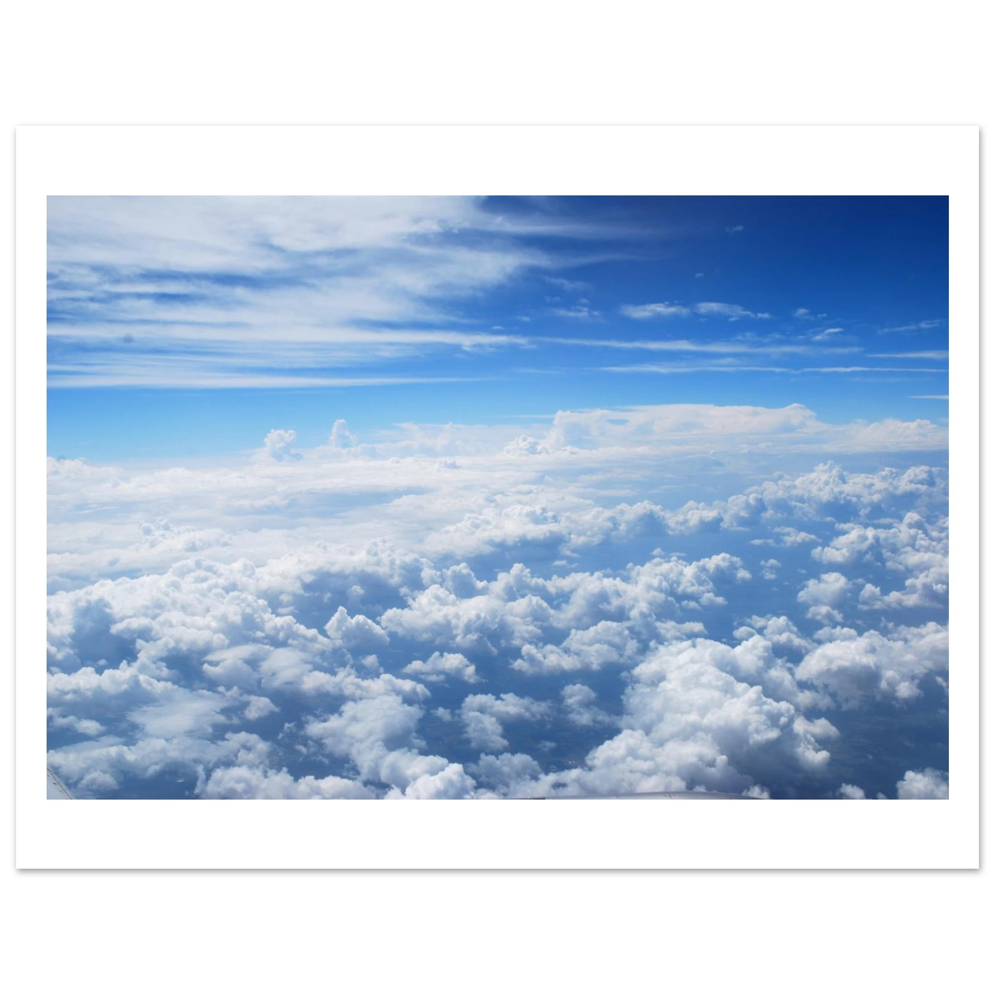 This is the 8" x 12" Above the Clouds Print by Lee Hansheng Studios. The rectangular stunning photograph of clouds captured from 36,000 feet above the earth, a breathtaking perspective that showcases the soft, ethereal beauty of the horizon. It is bright blue, dark blue, fluffs and streaks of clouds on the image. It has a white border and against a black and white background. 