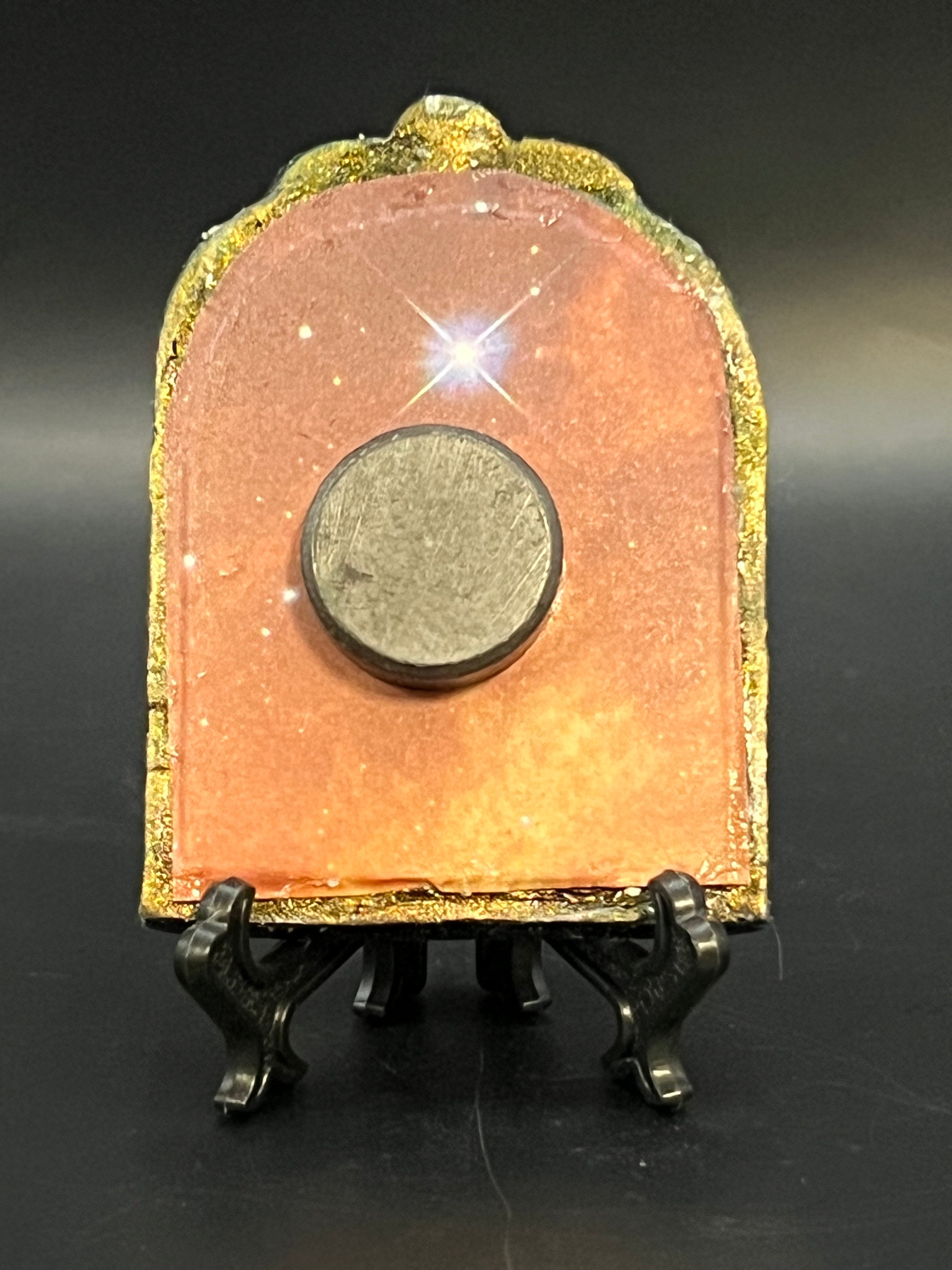 This is the handmade Alignment Miniature Painting by Chris Foster Design. This is showcasing the back. Decorative piece with a colorful, orange/yellow design with glistening stars on a stand against a dark background. There is a magnet and against a miniature picture frame.