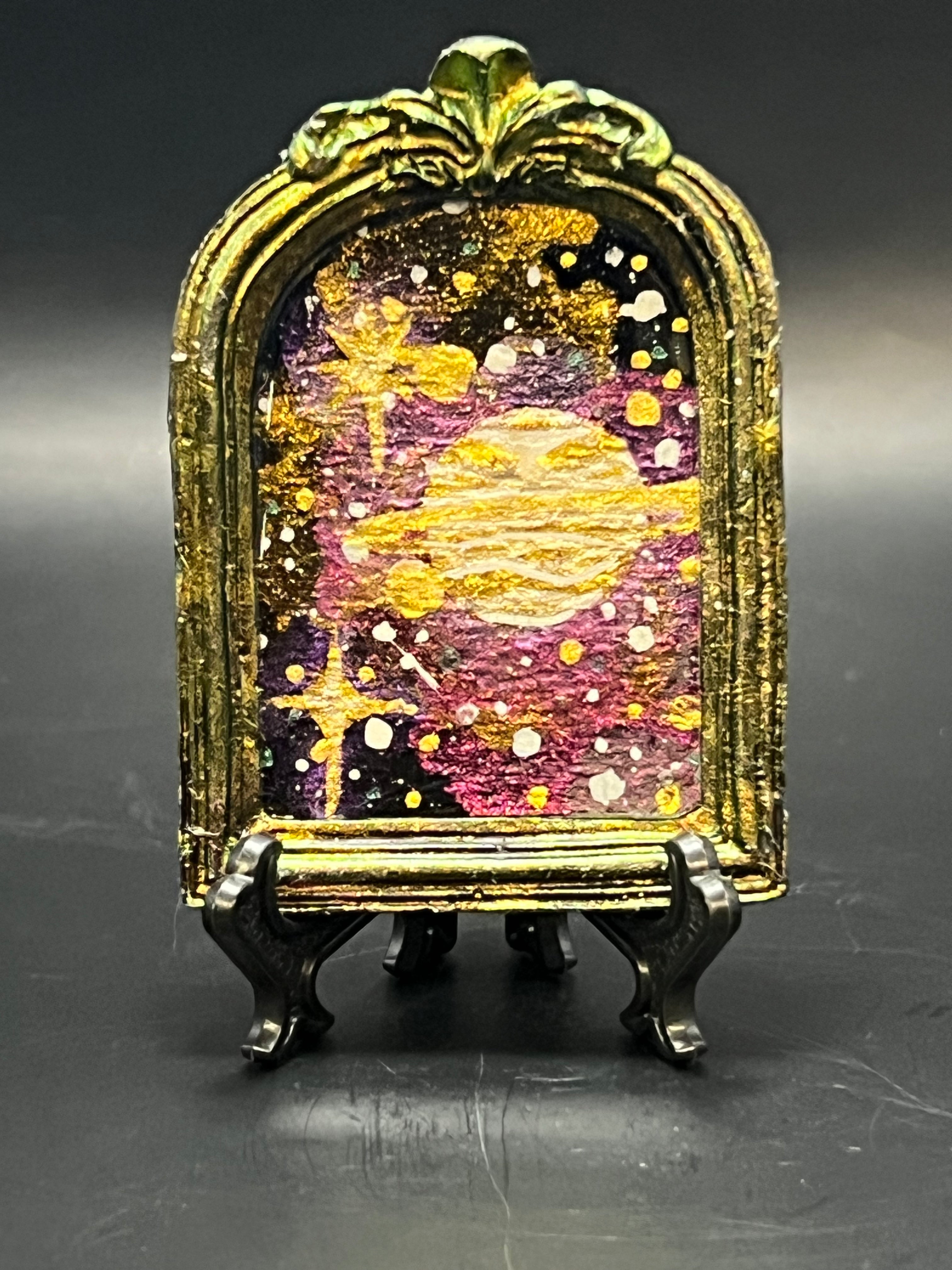 This is the handmade Alignment Miniature Painting by Chris Foster Design. This is showcasing the front. This is a decorative piece with a colorful, celestial design, orange/yellow with glistening stars on a stand against a dark background. There is a magnet and against a miniature picture frame.