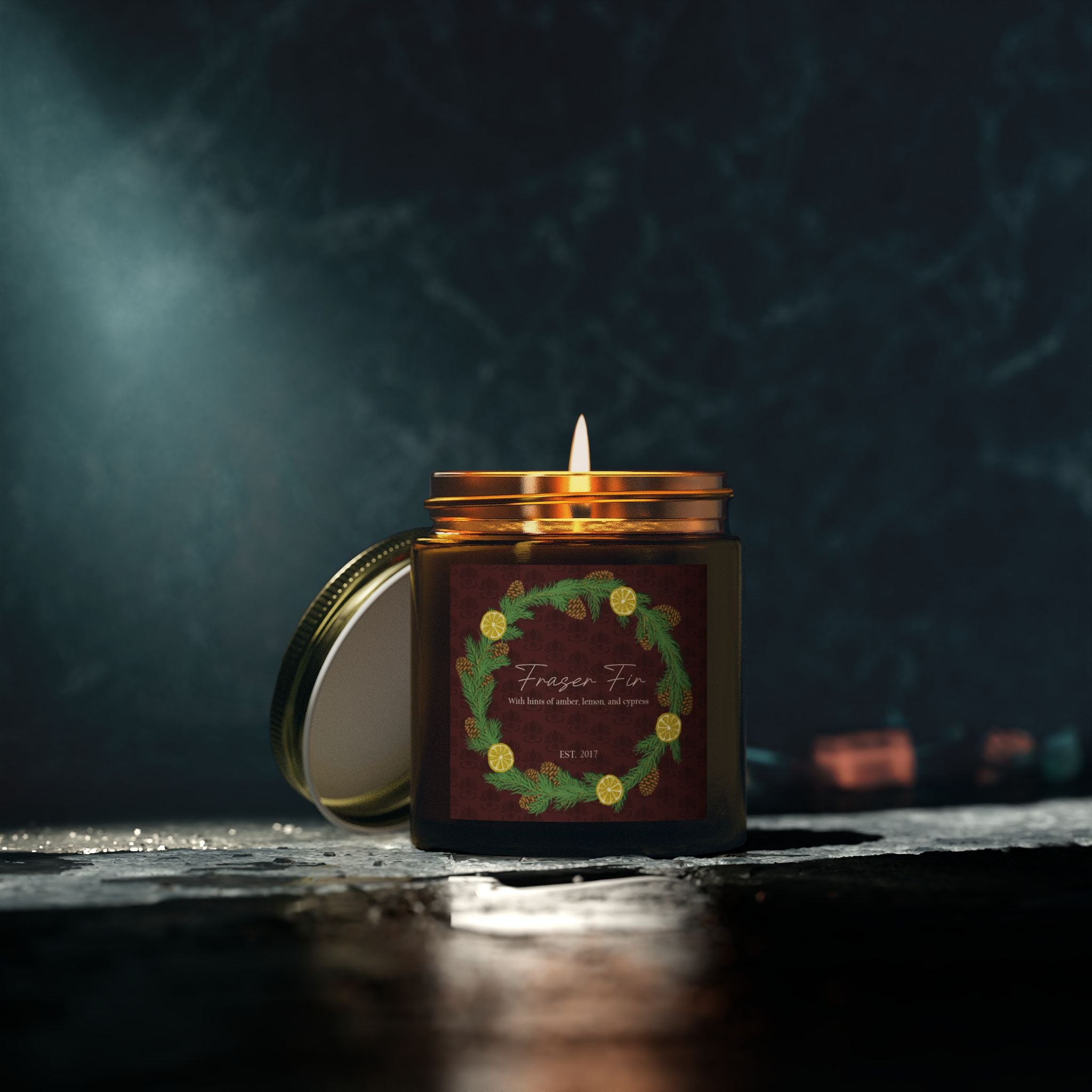 This is the 4oz Amber Fraser Fir Candle from Chris Foster Design. This is a photo from the front with the screw on top to the left and behind it. The candle is in a glass cylinder container that is tinted amber. The label is brown with a delicate pattern and has a lemon and pinecone wreath surround the name of the candle. The candle is on a black surface and a marble dark aqua background with some leaf decor in the back right.