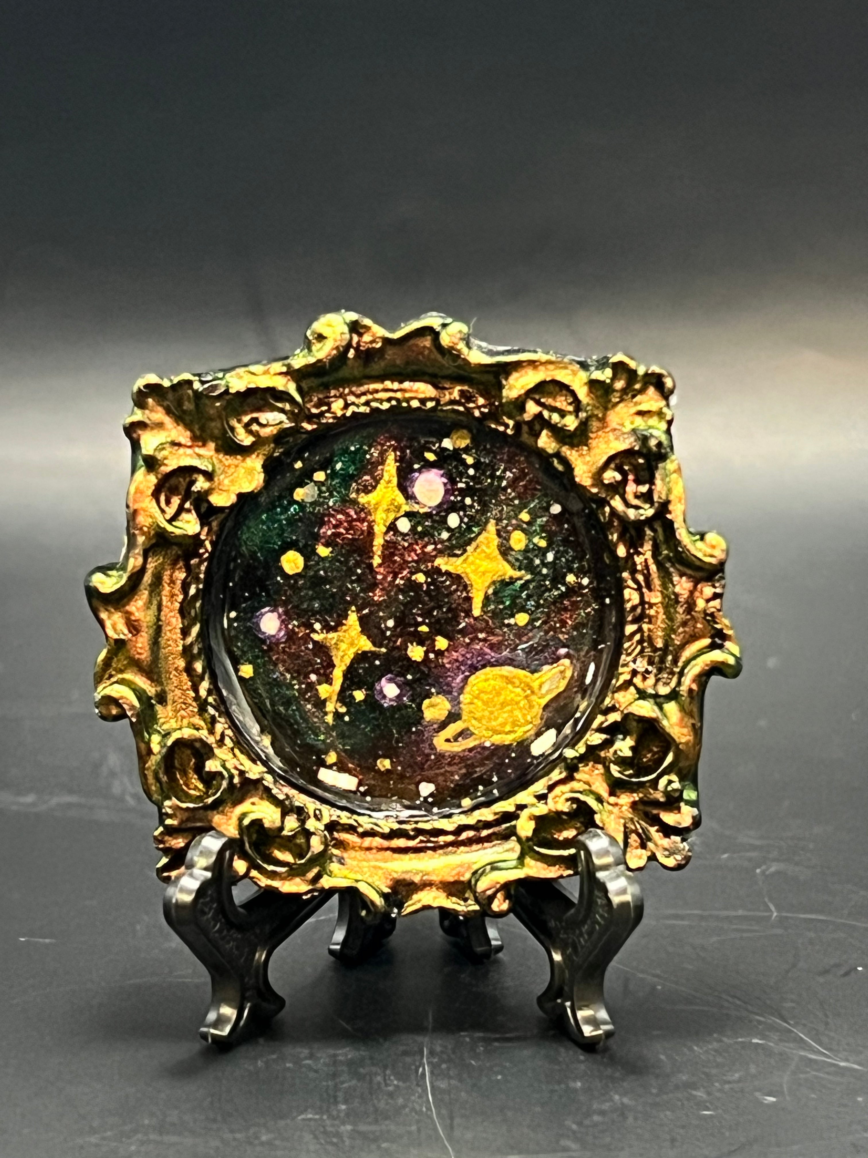 This is the handmade Ascension Miniature Painting by Chris Foster Design. This is showcasing the front. This is a decorative piece with a gold oval frame, starry night design including yellow stars, sparkling green, red, black and white smaller stars on a dark background. There is a magnet and against a miniature picture frame.