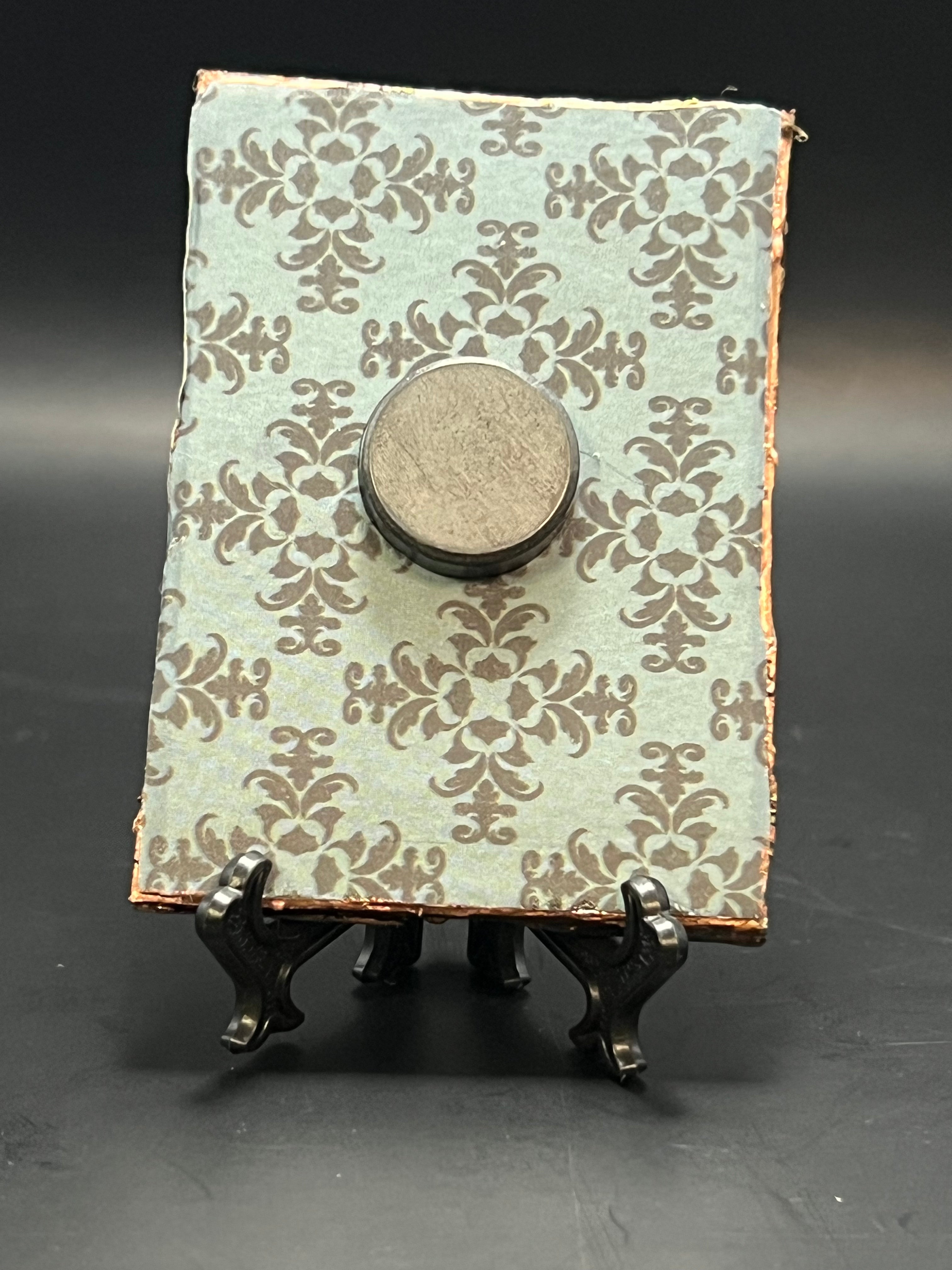 This is the handmade Assorted Beetles Miniature Painting by Chris Foster Design. This is showcasing the back. This is a small rectangular frame and the decorative item with a intricate pattern that is black on a blue background. There is a magnet and against a miniature picture frame.
