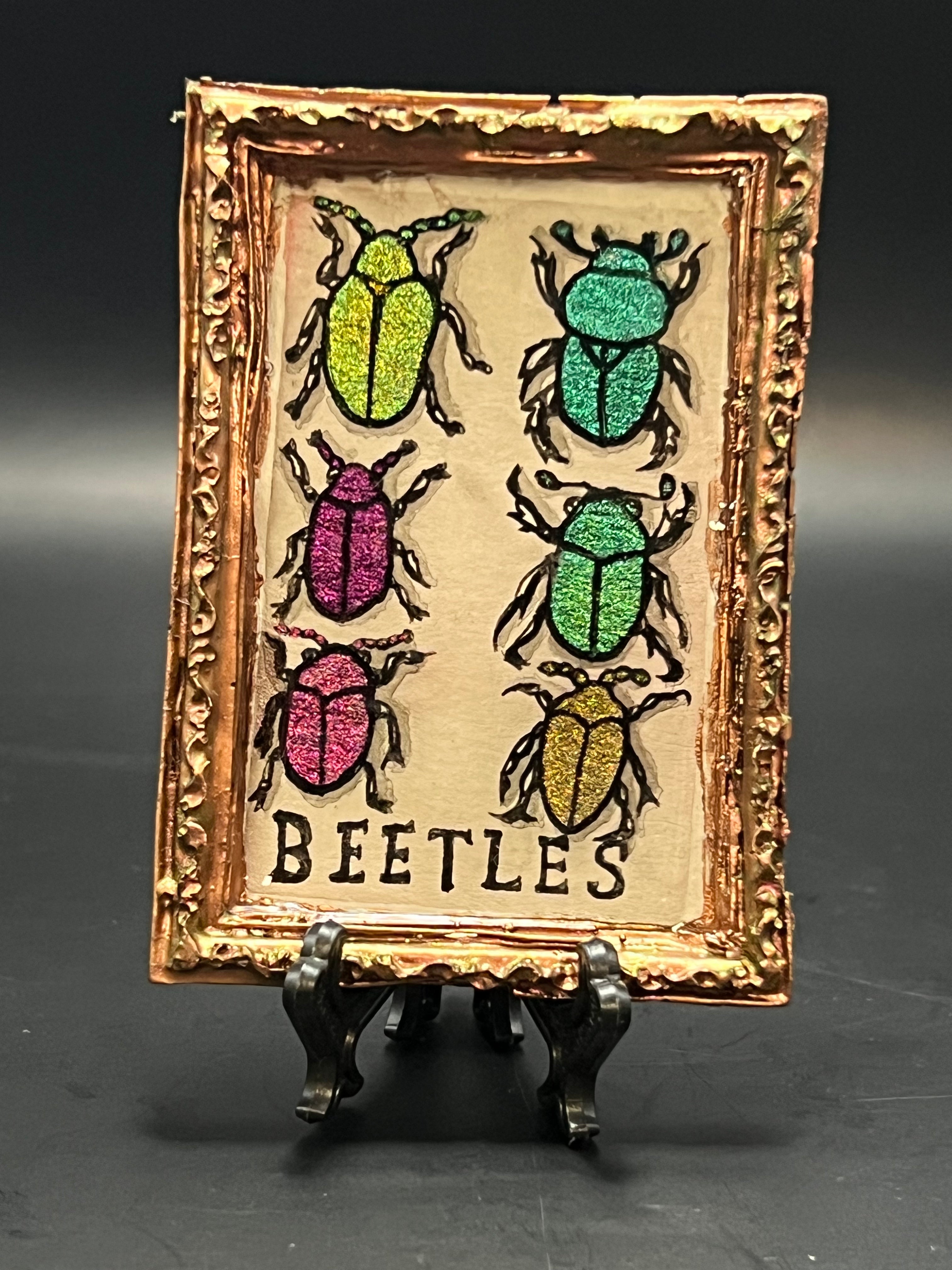 This is the Assorted Beetles handmade miniature painting by Chris Foster Design. It has a gold rectangular frame with illustrations of beetles in different shimmery colors of yellow, red, maroon, green, light green and gold on a tan background. The words, "BEETLES" are on the bottom of the painting. The frame is sitting on a stand.