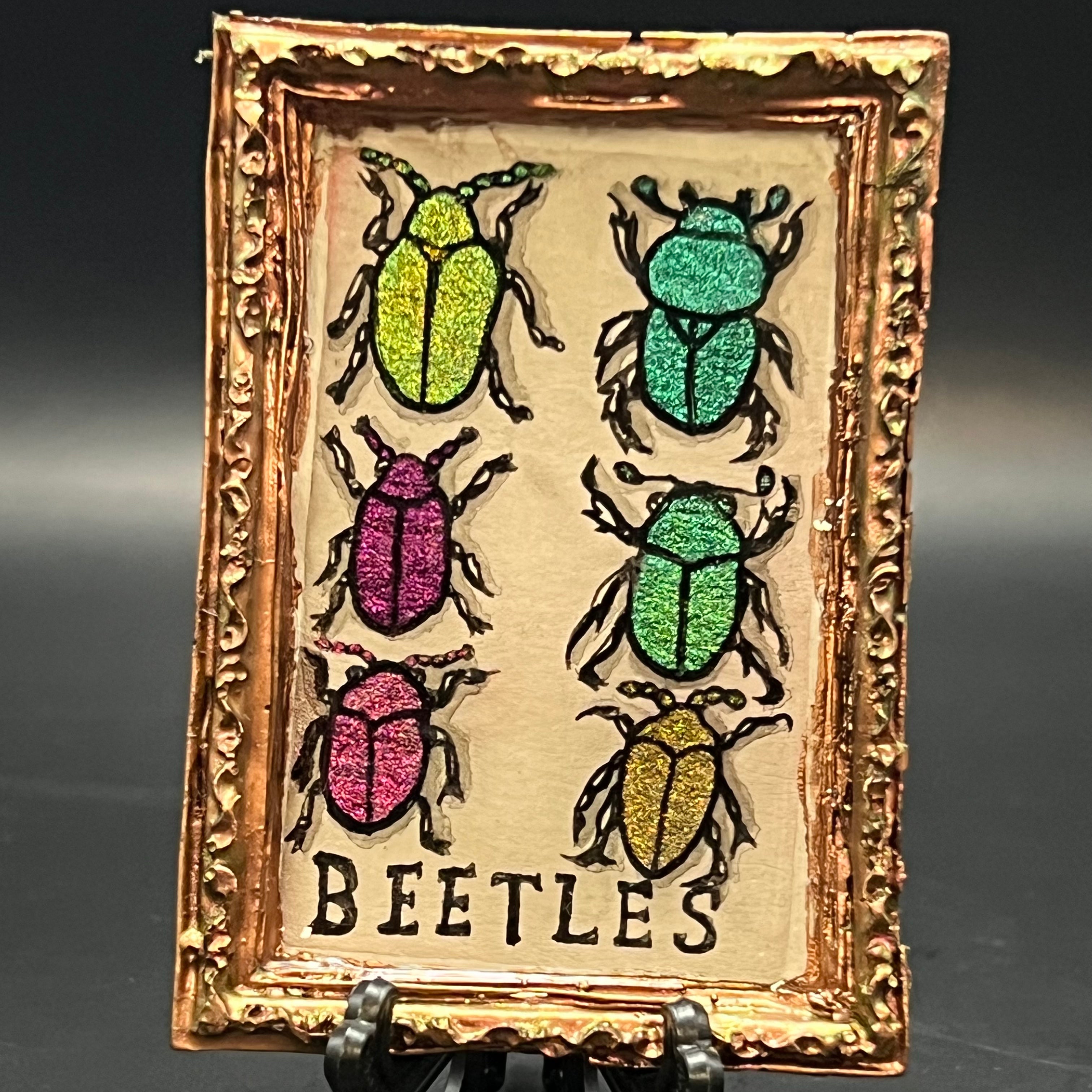 This is the Assorted Beetles Mini Painting by Chris Foster Design. This is showcasing the front. Large rose gold rectangle frame. Six assorted beetles on a light grey backround painted with color shift water colors. Hand lettered type BEETLES at the bottom. It is on a black stand and against a dark background.