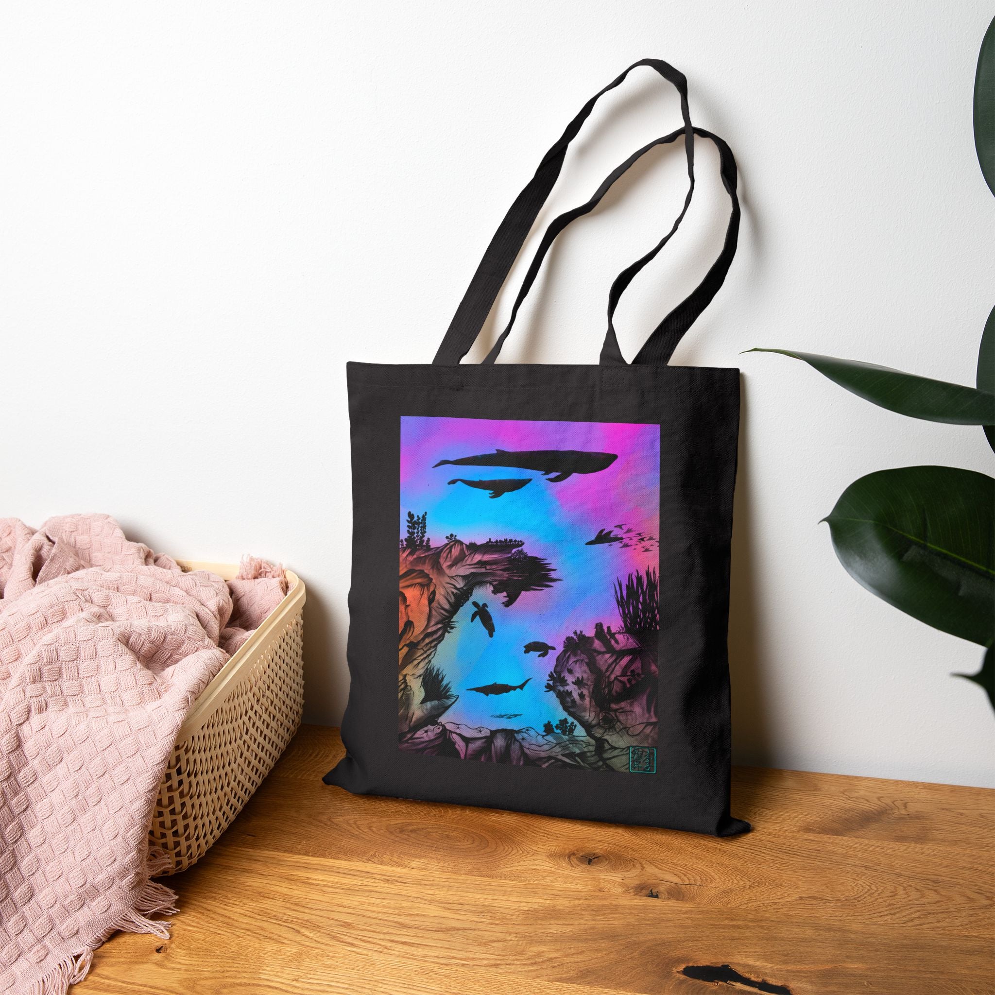 This is the Black Into the Abyss Canvas Tote by Lee Hansheng Studios.The illustration is underwater seascape with aquatic plants on a marbled background.There are two blue whales,1 goblin shark,school of eagle rays,two sea turtles and a school of fish.On the bottom right corner where the front patch of the backpack there is the studio logo printed in black outlined in blue.The colors are pink,purple,blue and red/orange.The tote is on a wooden surface with a pink blanket to the left and a plant to the right.
