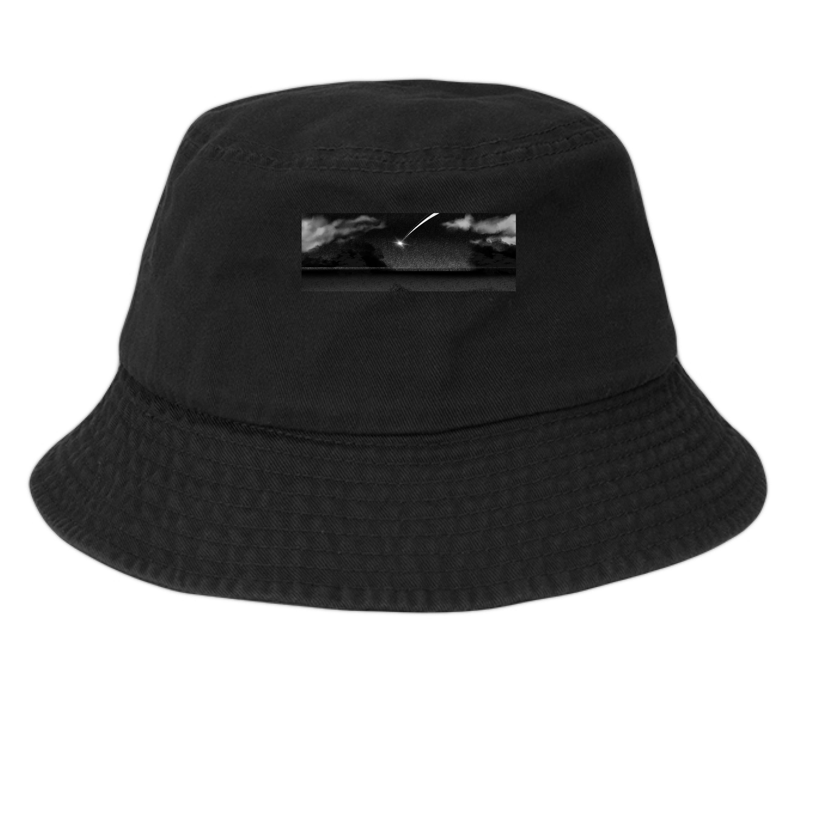 This is the Black Star Fall Bucket Hat by Lee Hansheng Studios. This is showcasing the hat from the front. The illustration is a meteor streaking through the sky. It is a moody piece that as gradients of black and grey, a little pixelated with white clouds in the forefront. The meteor is also white. It is a smaller rectangular than the cap. It is against a white background.