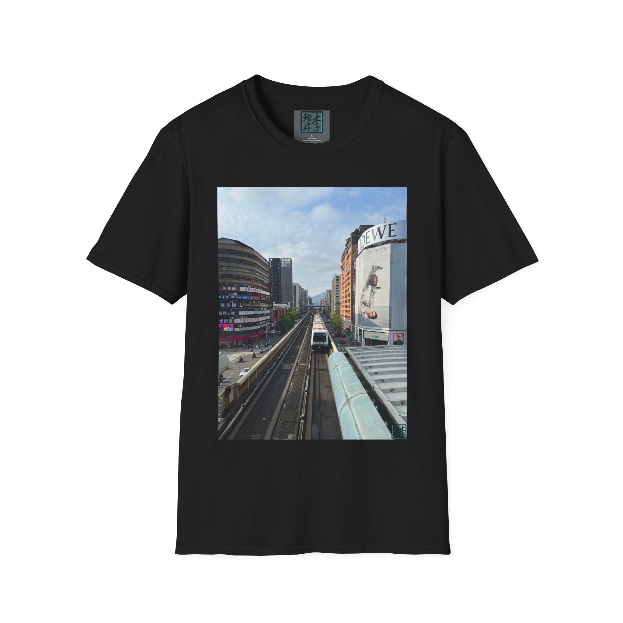 This is the Black Taipei MRT Day Softstyle T-Shirt by Lee Hansheng Studios. It is an image of the MRT train gliding along the dark road with tall buildings to the left and right. The builing to the left is white and behind it is a brick building. to the right is a glass bulding with different advertisements on it. There are various colors under a blue cloudy sky. It is against a white background. 