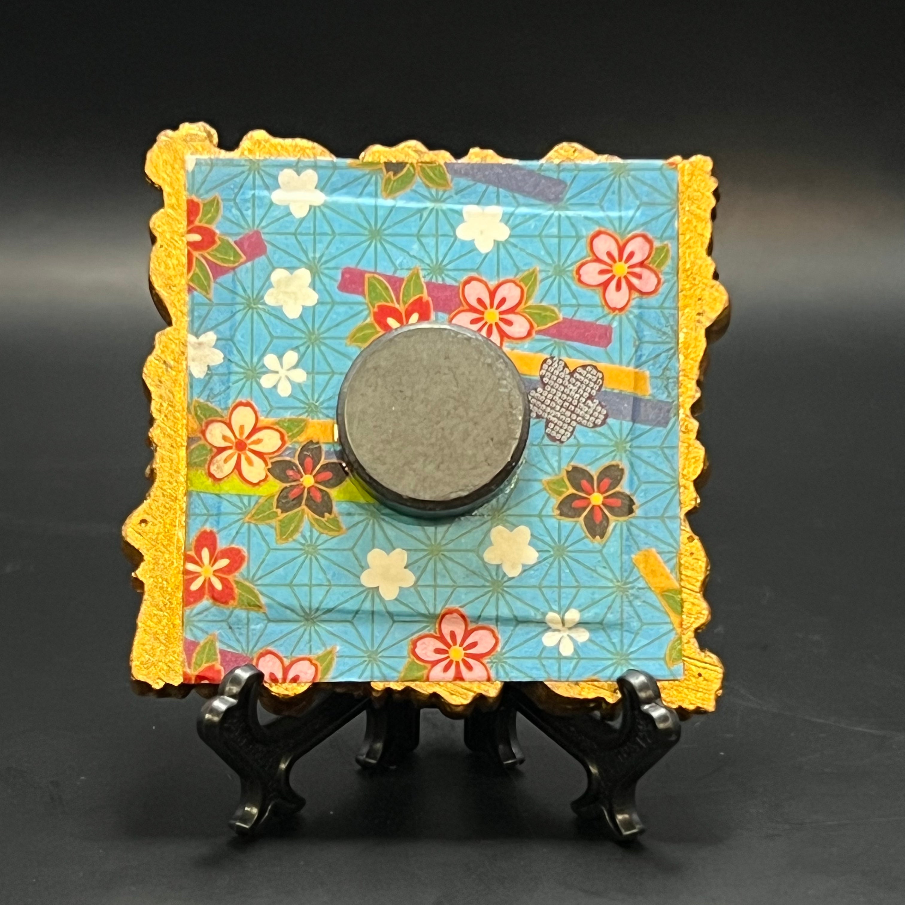 This is the Blue Plaid Stars Mini Painting by Chris Foster Design. This is showcasing the back. A decorative frame with floral pattern in white, yellow, red and black with a blue background. The painting is on a stand against a black background.