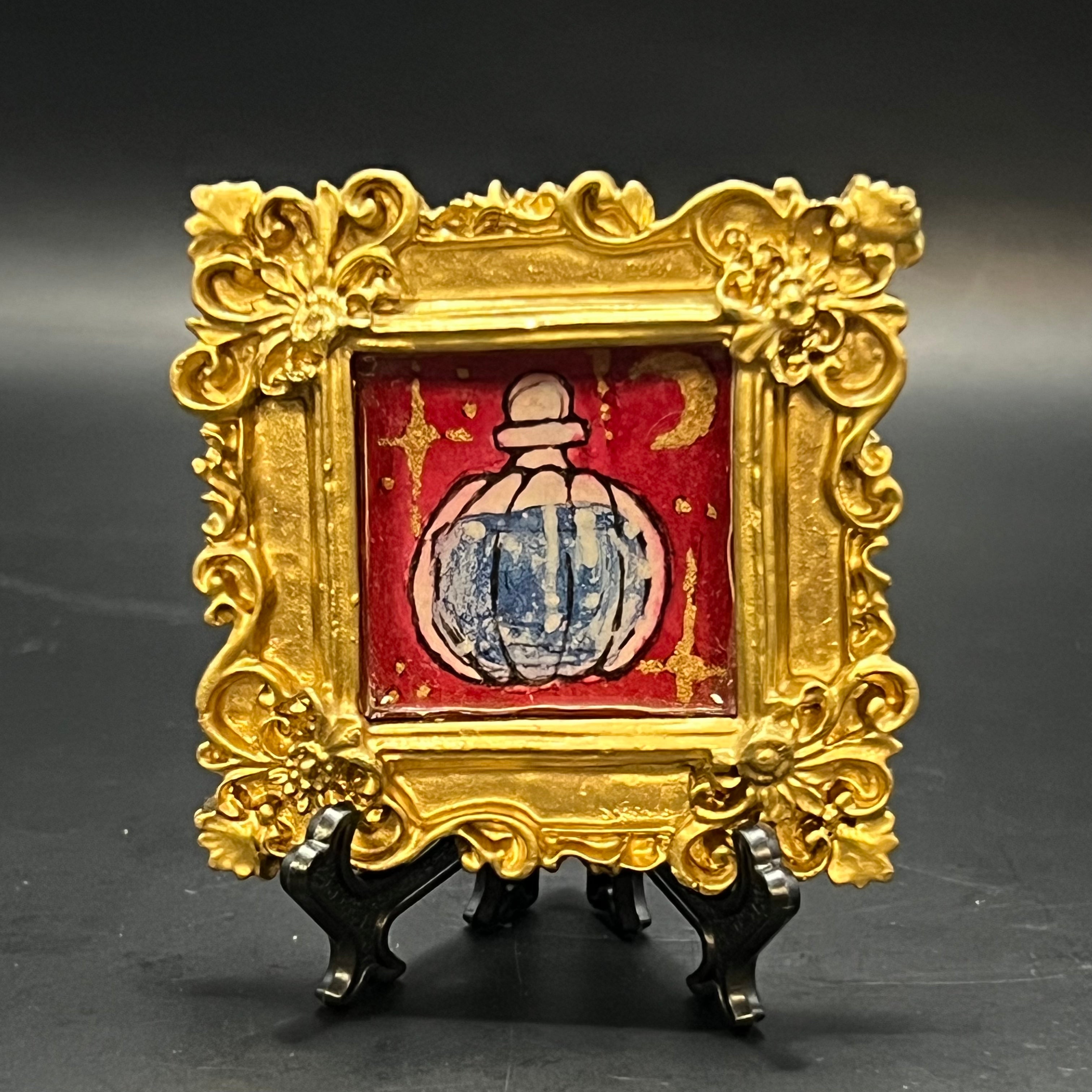This is the Blue Potion Mini Painting by Chris Foster Design. There is a decorative gold frame with a red background with glistening yellow stars and moon. In the center is a glass potion bottle with glittery blue liquid inside. It's against a dark background.
