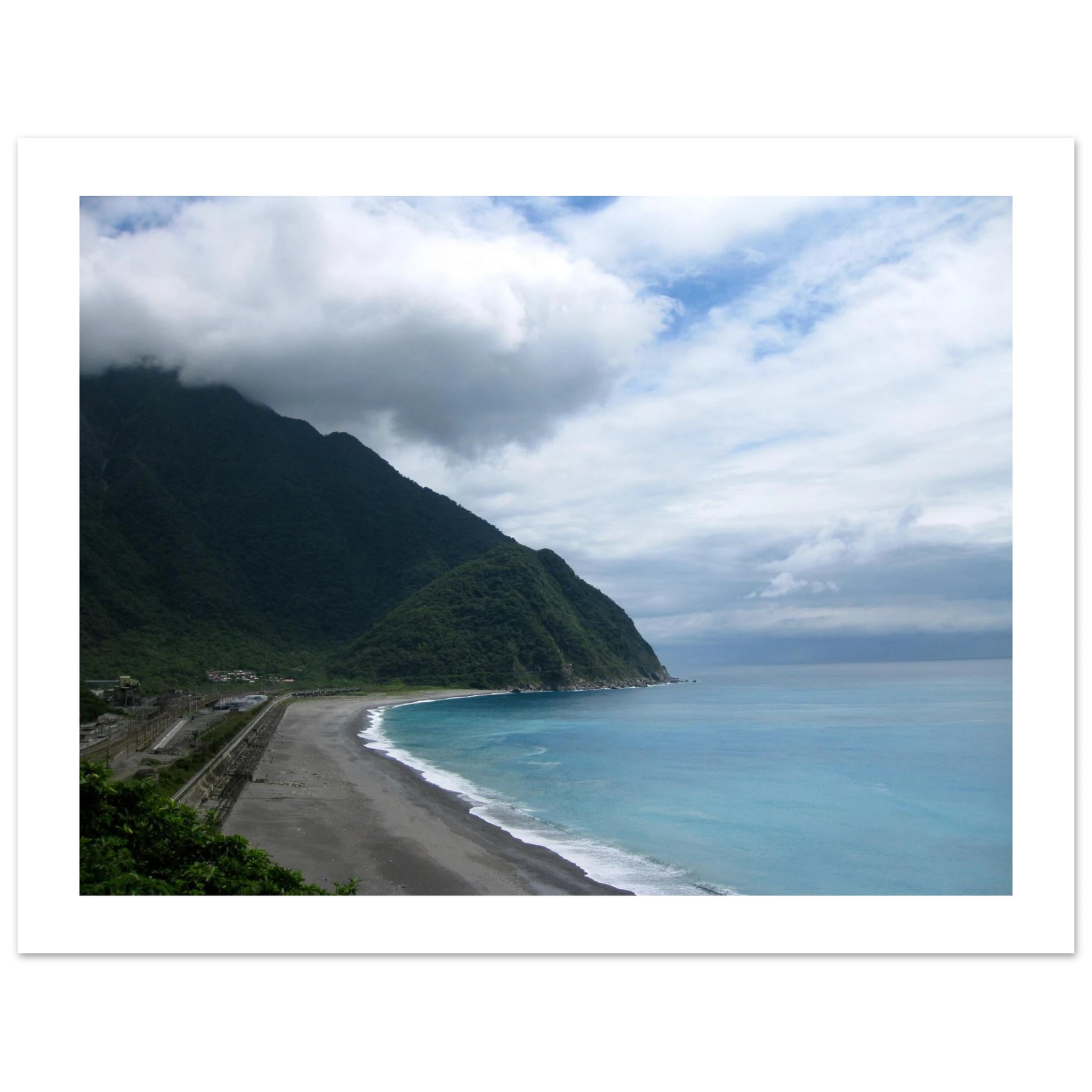This is the 8" x 10" By Mountains and Sea Print by Lee Hansheng Studios. It is of a famous coast of Taroko National Park in Taiwan. The coastal scene with a beach, dark sand, ocean, and mountains under a cloudy sky. To the left are some houses. There is a white border around the print. 