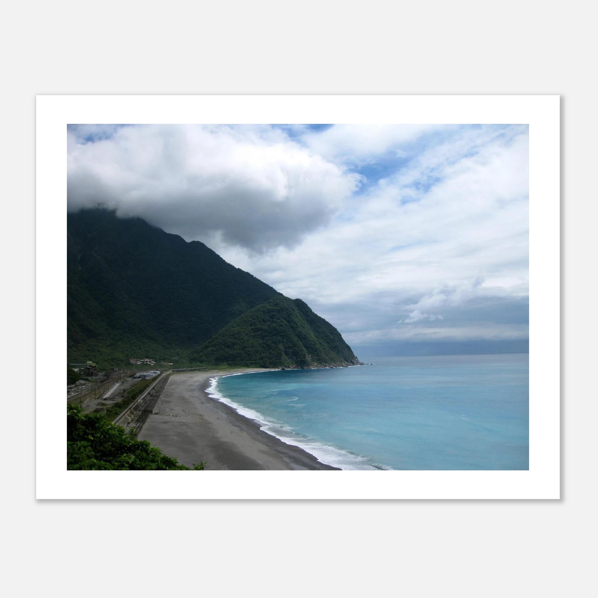 This is the 8" x 10" By Mountains and Sea Print by Lee Hansheng Studios. It is of a famous coast of Taroko National Park in Taiwan. The coastal scene with a beach, dark sand, ocean, and mountains under a cloudy sky. To the left are some houses. There is a white border around the print and against a white background. 