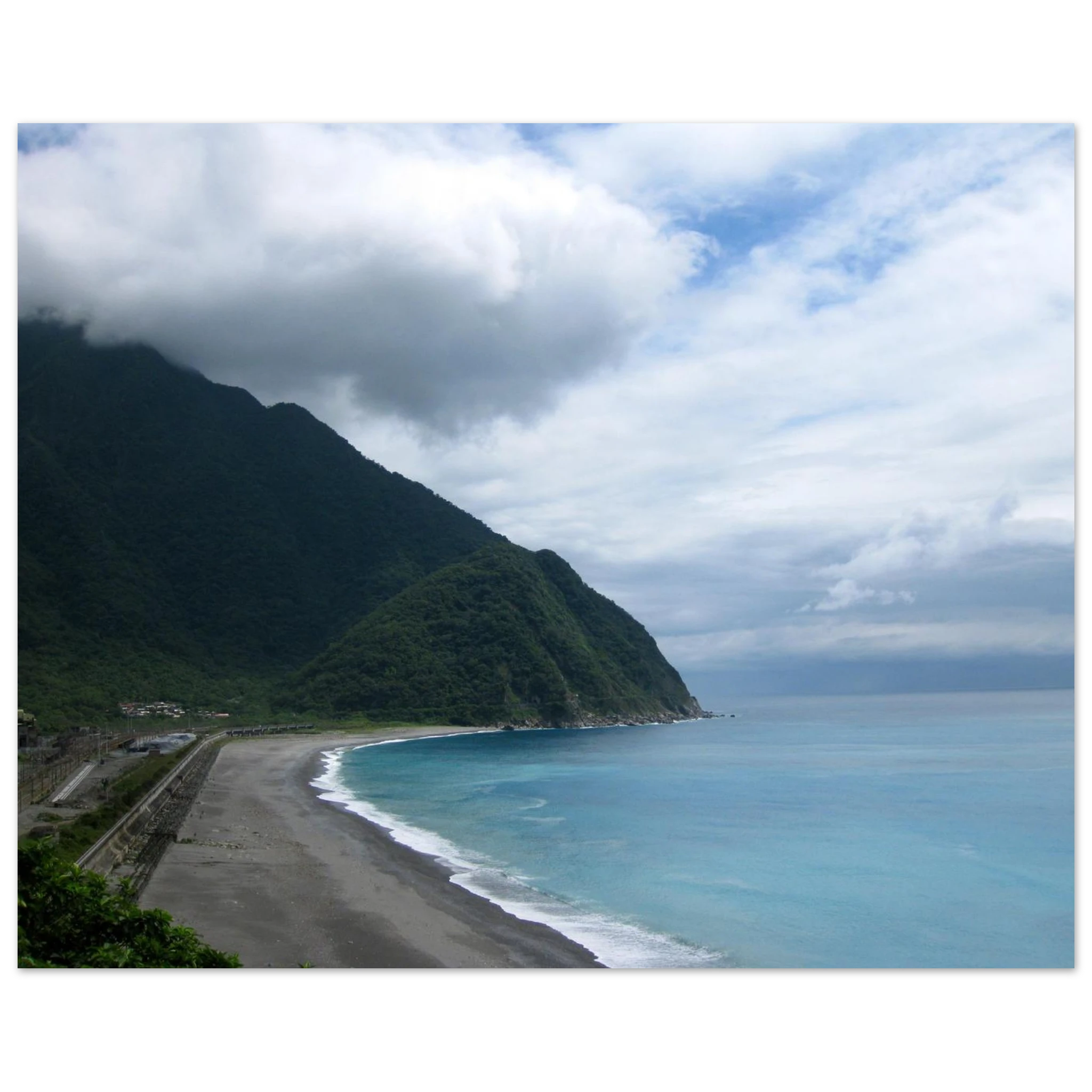This is the 8" x 12" By Mountains and Sea Print by Lee Hansheng Studios. It is of a famous coast of Taroko National Park in Taiwan. The coastal scene with a beach, dark sand, ocean, and mountains under a cloudy sky. To the left are some houses. 