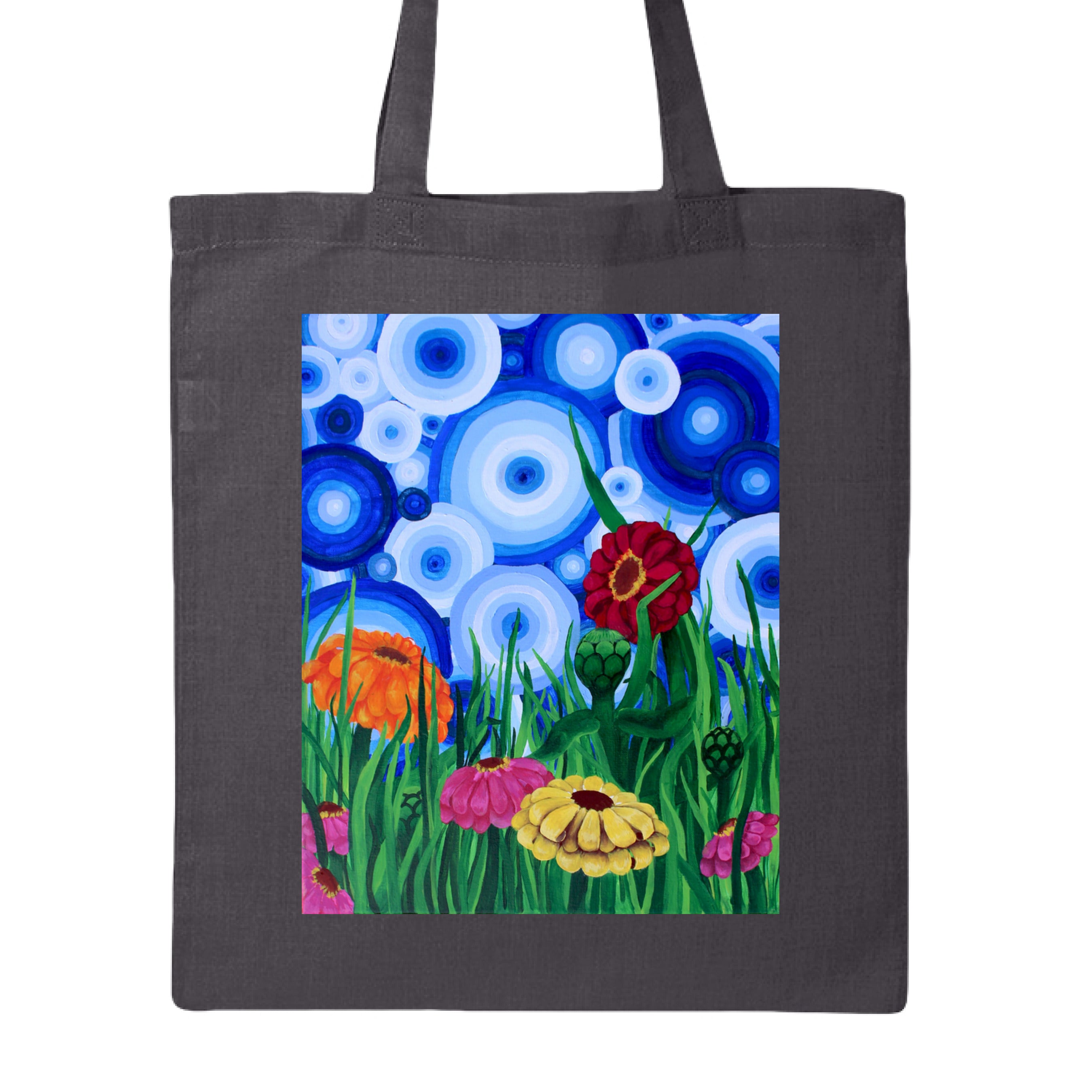 This is the Charcoal Zinnia 2 Canvas Tote by Storm Garden Studio. This showcases the front of the tote. The rectangular illustration are bold and color zinnias like red, yellow and orange across tall green grass. The background has many different circles in blue, dark blue and white. The tote is against a white background.