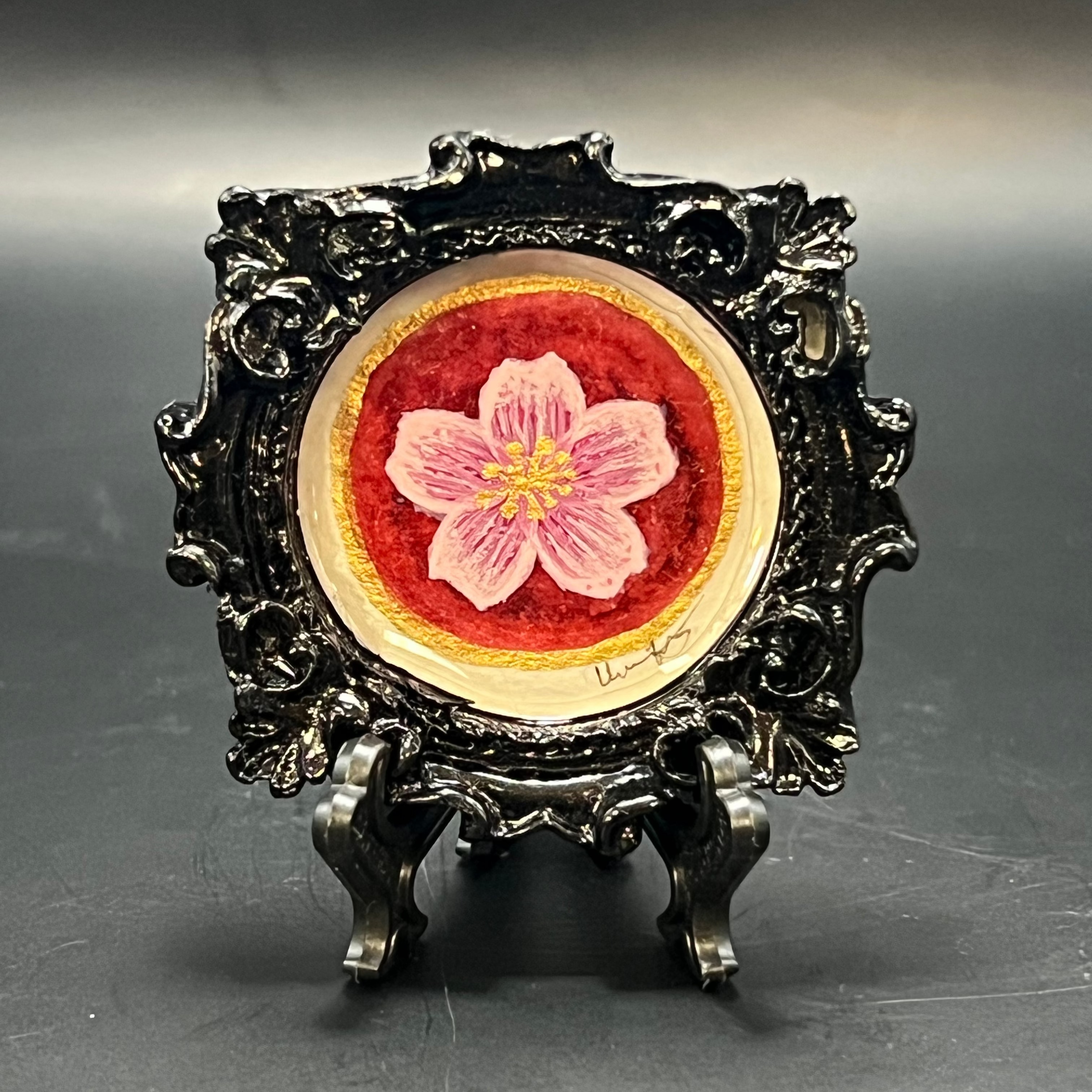This is the Cherry Blossom Bloom Mini Painting by Chris Foster Design. This is showcasing the front. Ornate Gloss black circle frame. Single cherry blossom paining in light pink with gold stamens, and deep red with gold glitter background. It against a dark background.