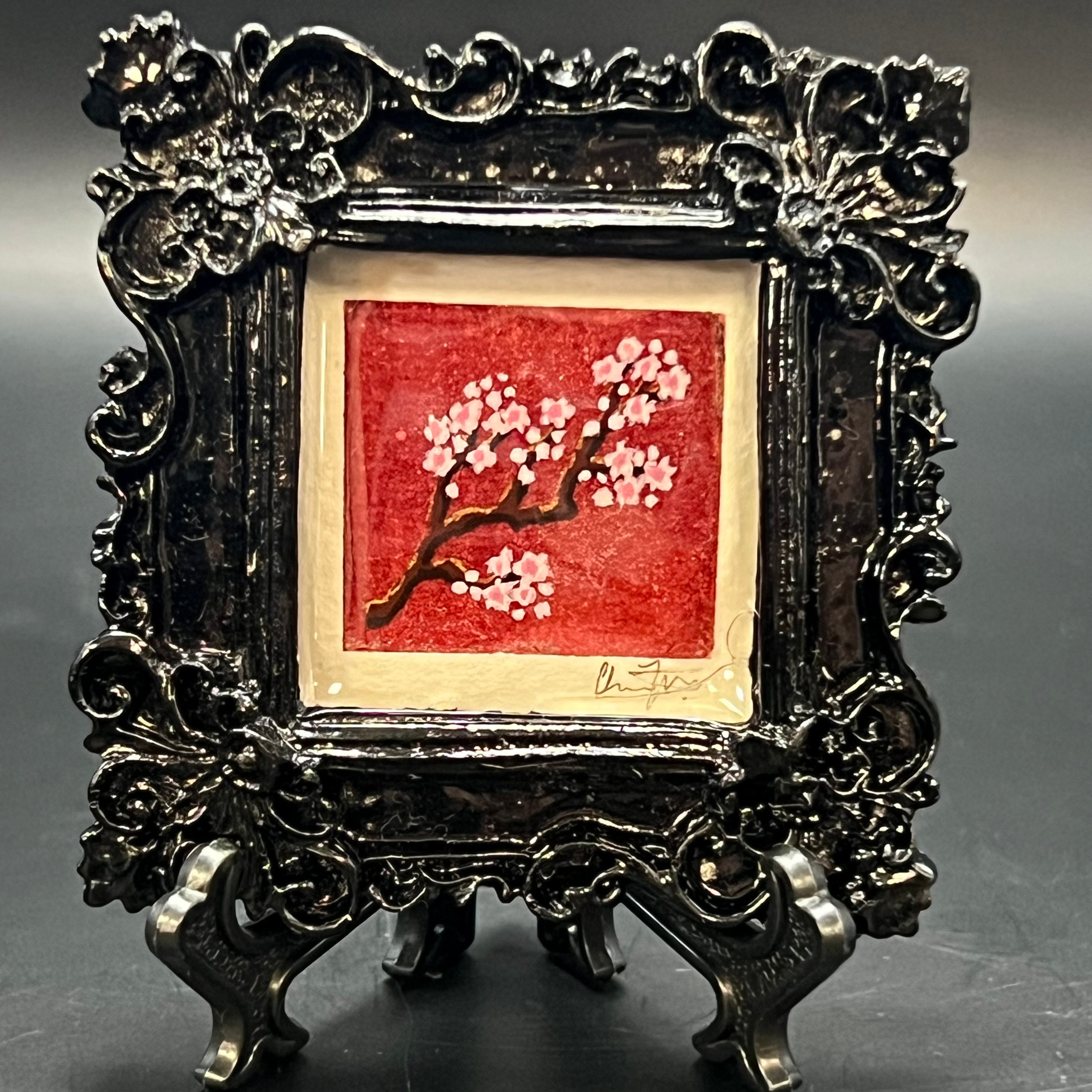 This is the Cherry Blossom Branch Square Mini Painting by Chris Foster Design. This is showcasing the front. Gloss black square ornate frame. Diagonal Cherry blossom branch with light pink flowers, and deep red back round with gold glitter wash. It is against a dark background. 