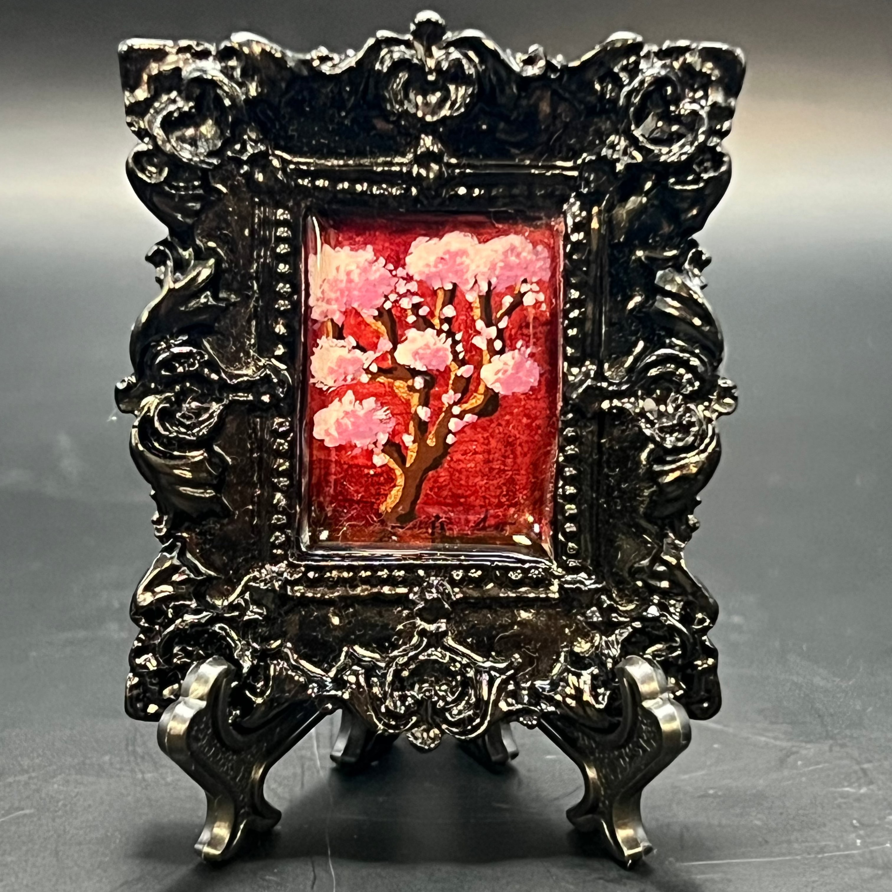 This is the Cherry Blossom Vertical Mini Painting by Chris Foster Design. This is showcasing the front of the painting. Gloss black rectangular ornate frame. Cherry Blossom branch in full bloom in light and dark pink with deep red and gold glitter background. It is against a dark background.