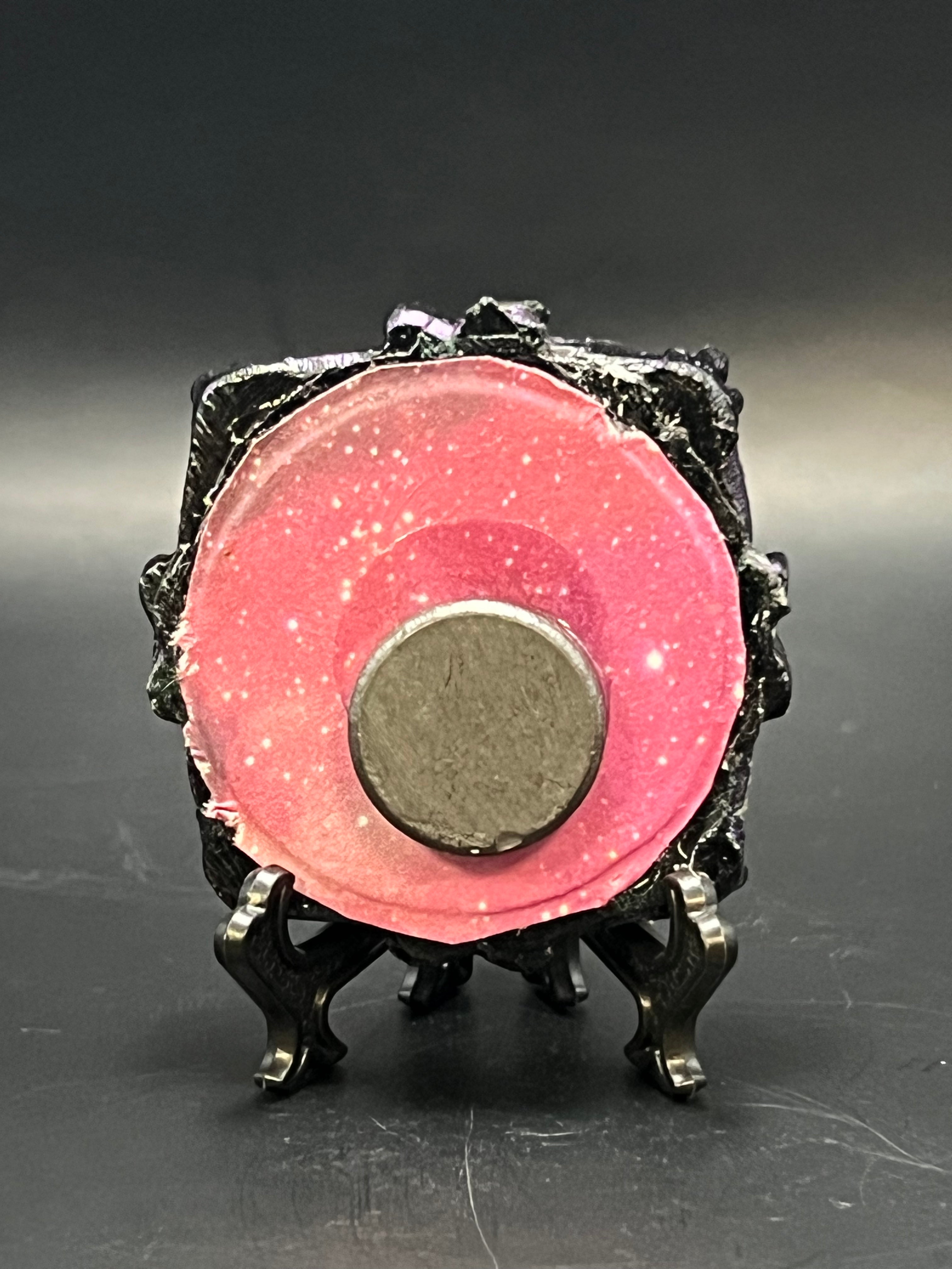 This is the handmade Clustered Crescent Miniature Painting by Chris Foster Design. This is showcasing the back.  This is a small circular frame and the decorative item with a sparkly mix background with pink and orange on a stand with yellow stars. There is a magnet and against a miniature picture frame.